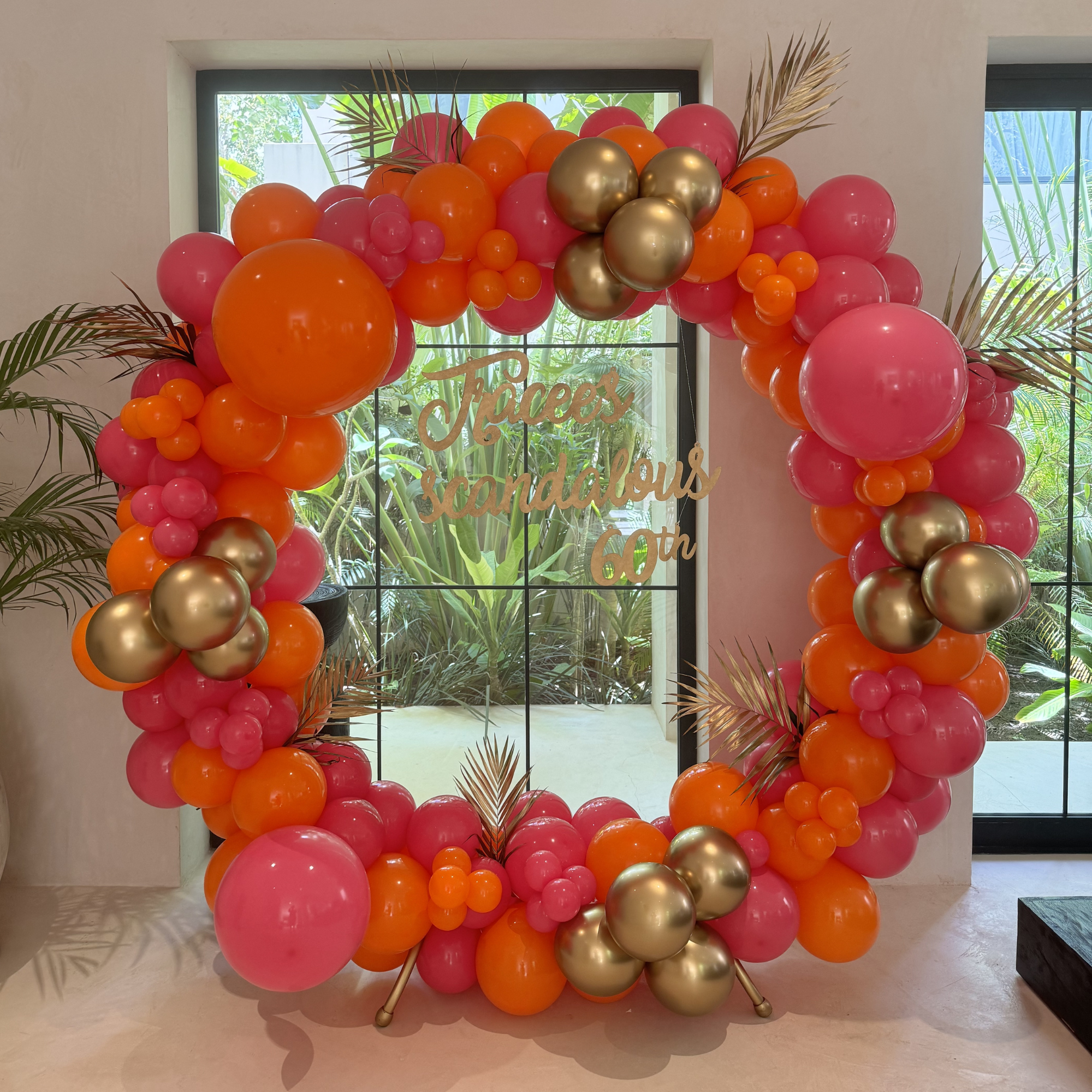 Decorative balloon arch with pink, orange, and gold balloons, accented with palm leaves, in front of window with foliage outside.