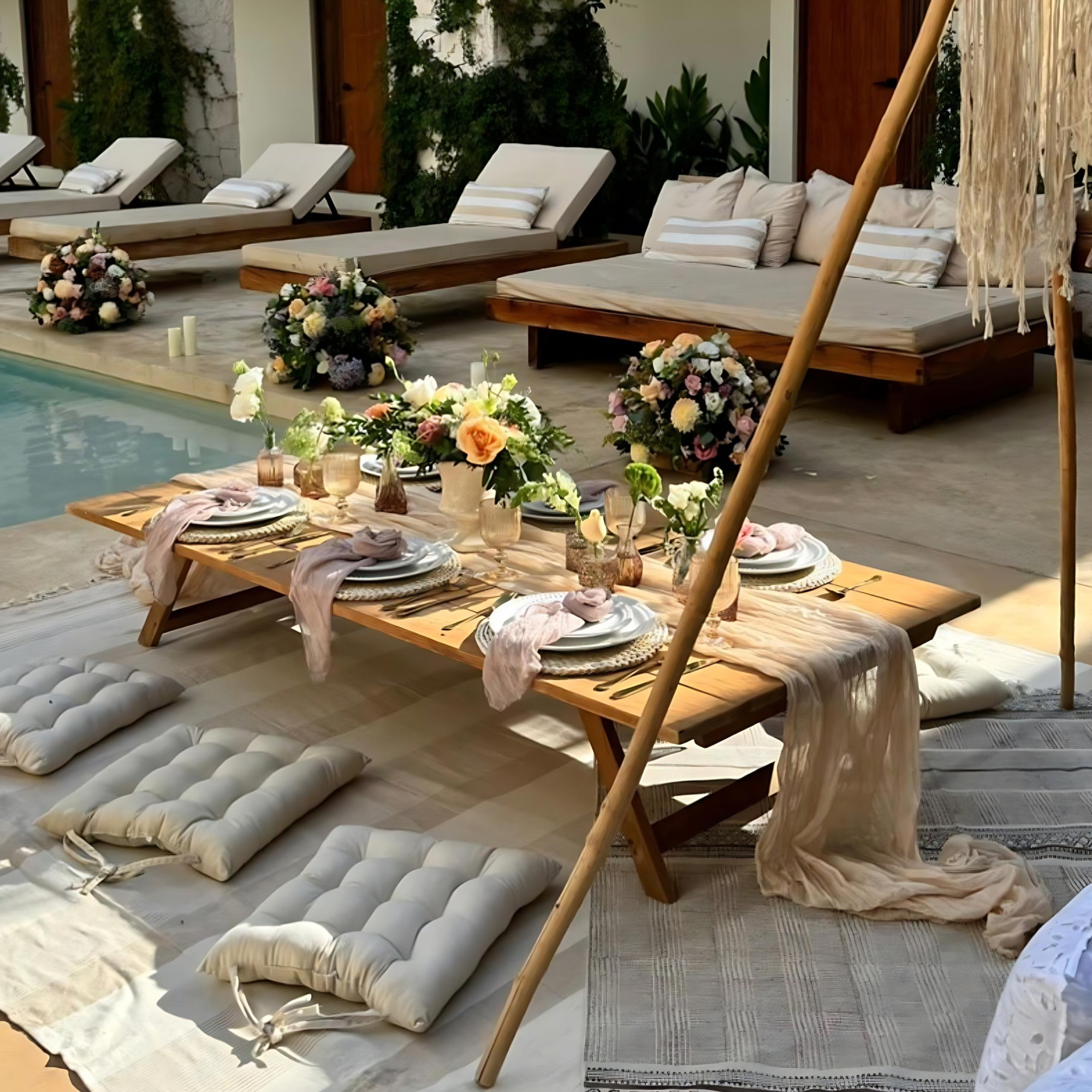 Indoor space decorated for a special occasion, with a low wooden table set for a meal, surrounded by woven baskets and cream-colored pillows, with floral arrangements and candles, and lounge chairs in the background.