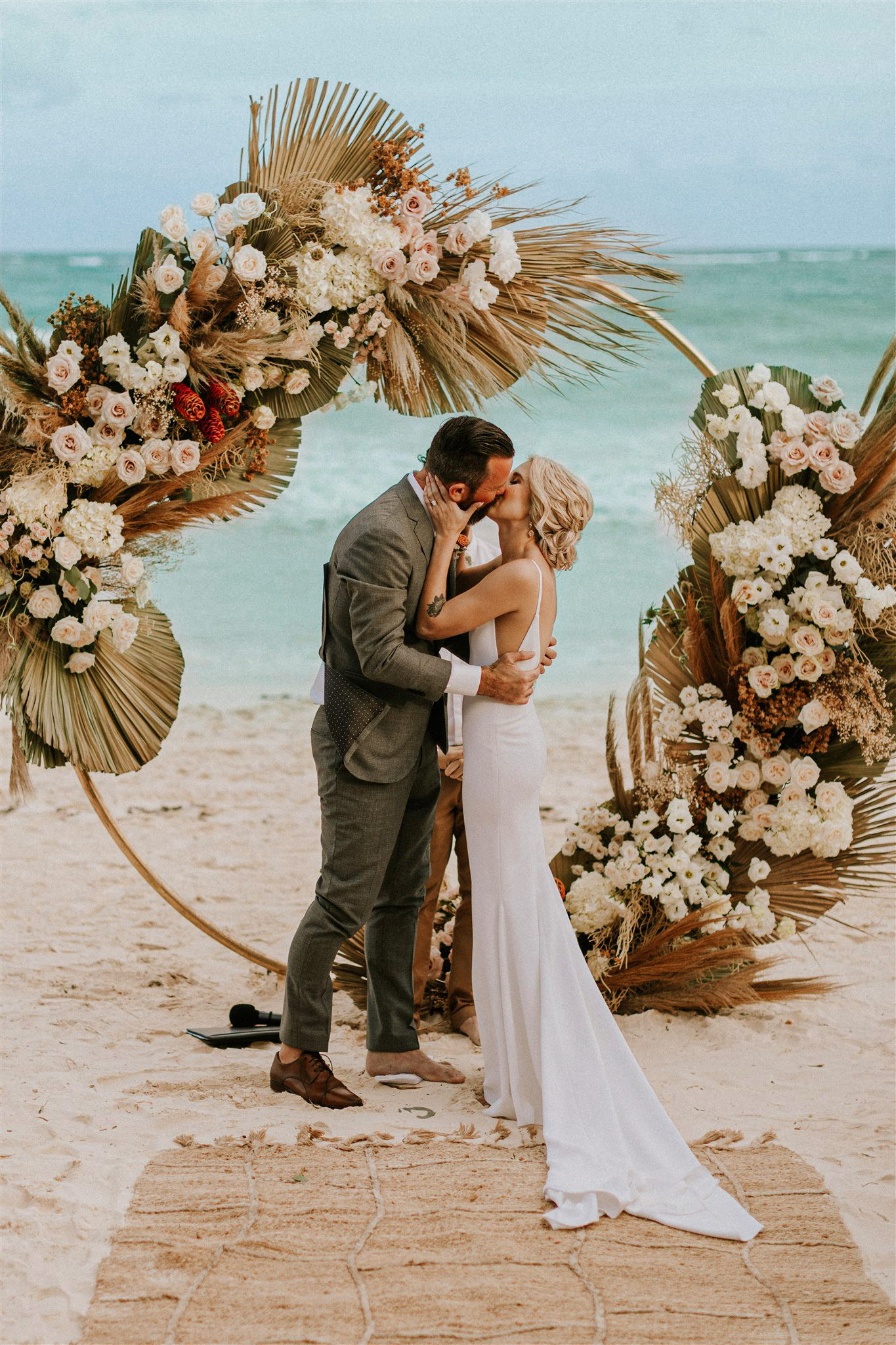 A couple kissing during a beach wedding ceremony, standing on a woven rug with a large floral and palm leaf arch behind them, ocean in the background.