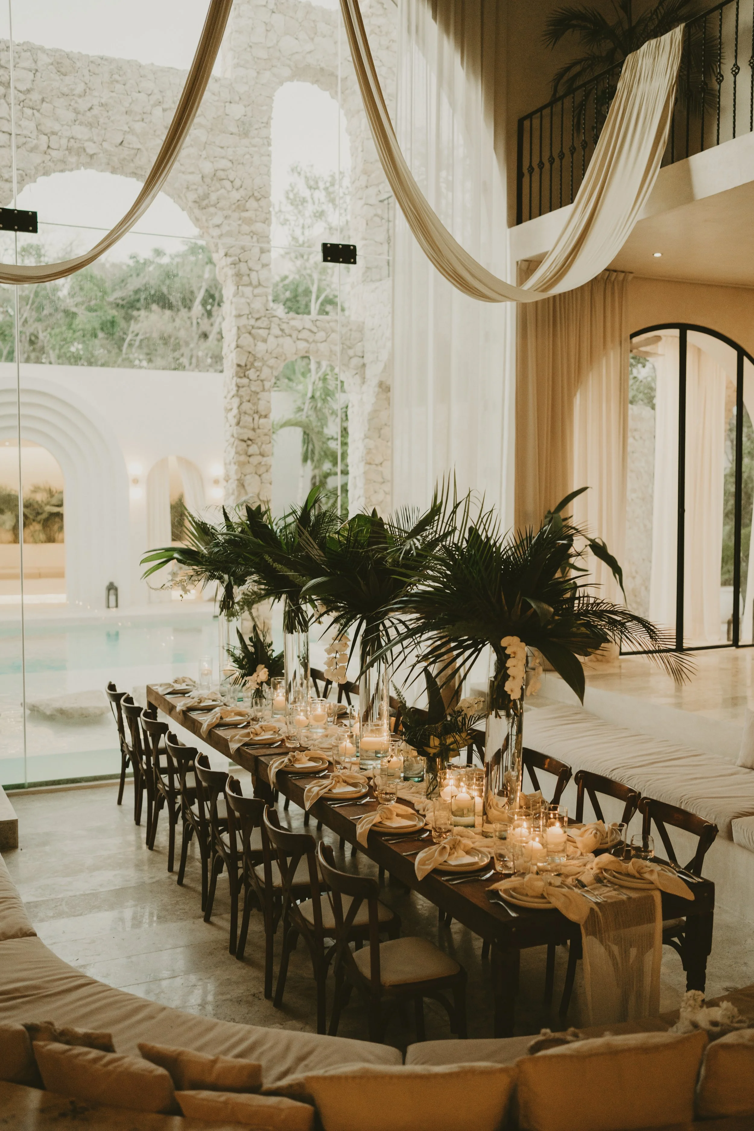 Elegant indoor dining area with a long table decorated with candles and floral arrangements, surrounded by dark wooden chairs, large plants, and a high ceiling with flowing white drapes. Large windows reveal a stone arch and outdoor scenery.
