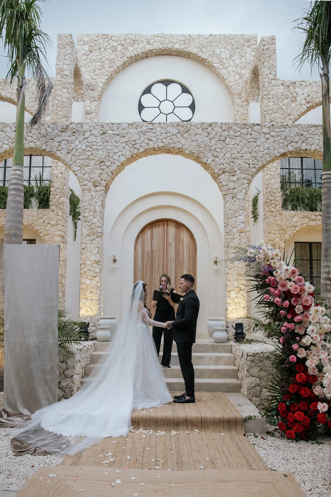A wedding ceremony taking place outdoors in front of a stone building with an arched wooden door, surrounded by palm trees and adorned with a large floral arrangement on the right. The bride and groom are holding hands, facing each other, with an off