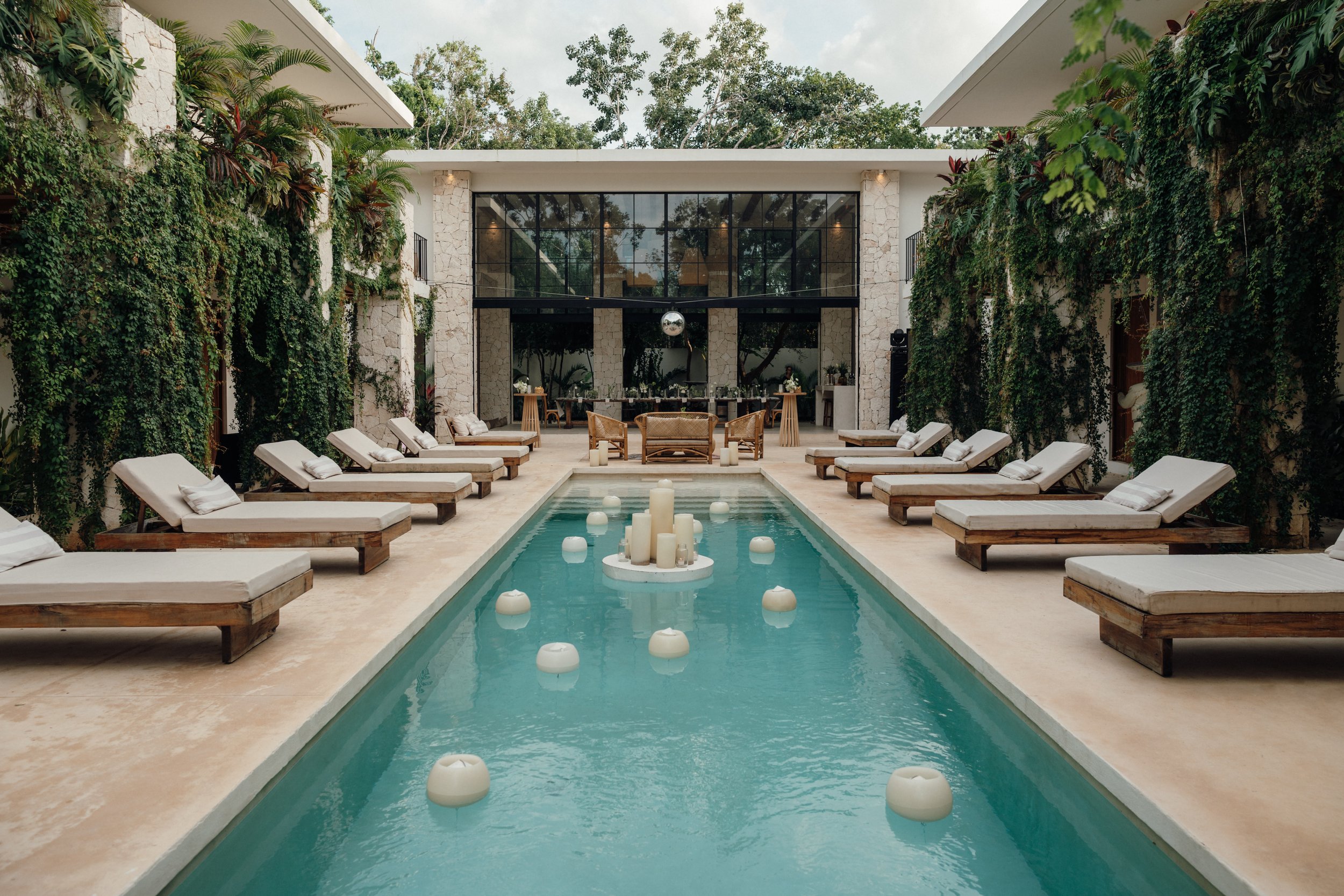 Outdoor lounge area with a rectangular pool, surrounded by lounge chairs with cushions, and a modern building with large glass windows and greenery.