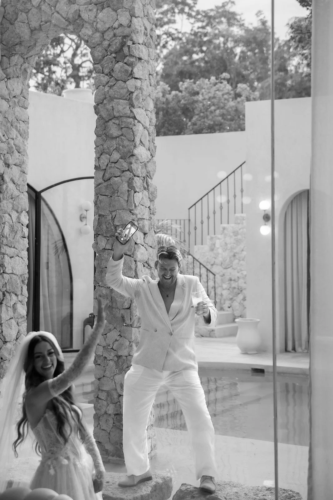 Man and woman celebrating outdoors, with the man holding a drink and a phone, and the woman reaching out to him, smiling. They are near stone structures, with palm trees and outdoor lighting visible.