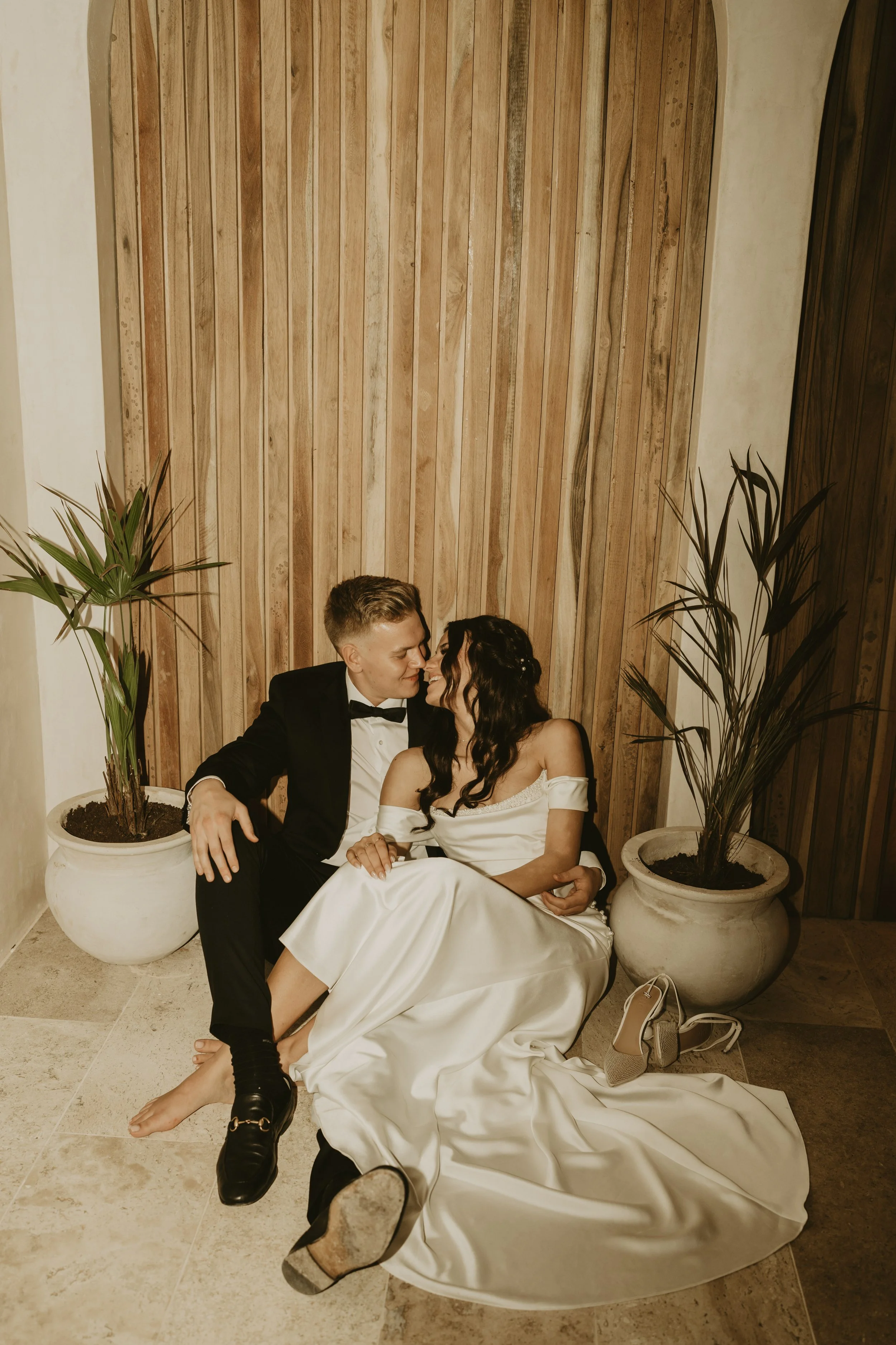 A newlywed couple sitting on the floor, sharing an intimate moment with their faces close together. The groom is wearing a black tuxedo with a bow tie, and the bride is dressed in a white wedding gown with off-shoulder sleeves. They are surrounded by