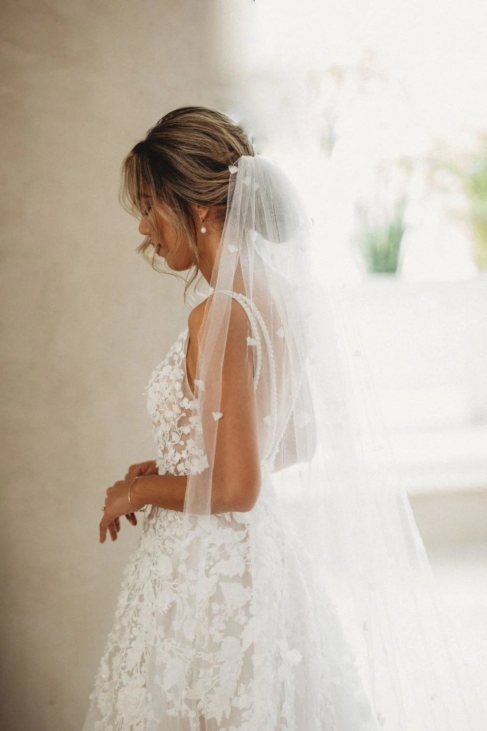 A bride in a white wedding dress with lace details, wearing a veil and pearl earrings, standing indoors with soft lighting.