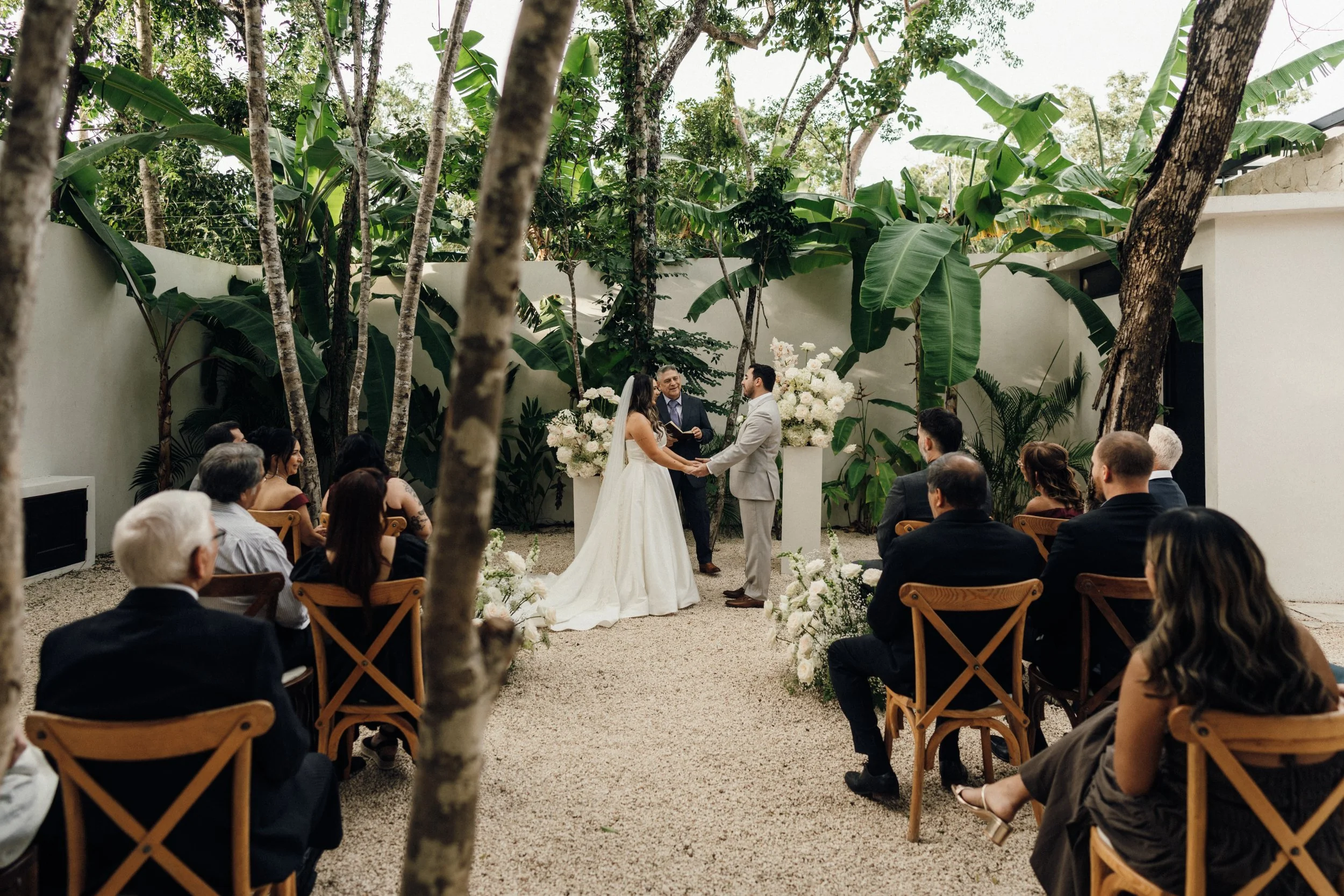 A wedding ceremony taking place outdoors in a garden-like setting with lush green trees and plants. The bride and groom are standing in front of an officiant, holding hands and exchanging vows. Guests are seated on wooden chairs watching the ceremony