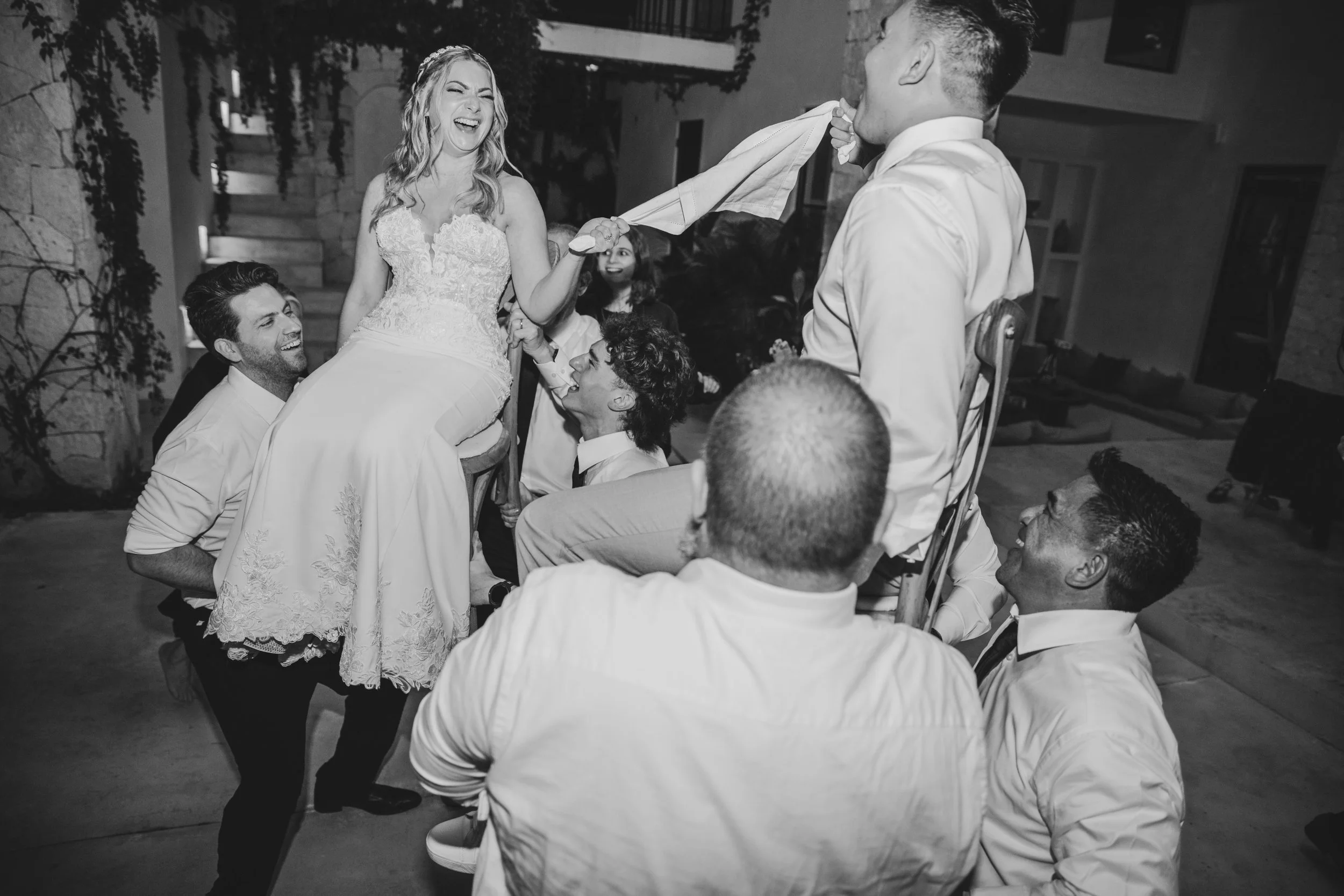 A woman in a wedding dress laughing and holding hands with a man, surrounded by friends at a celebration.