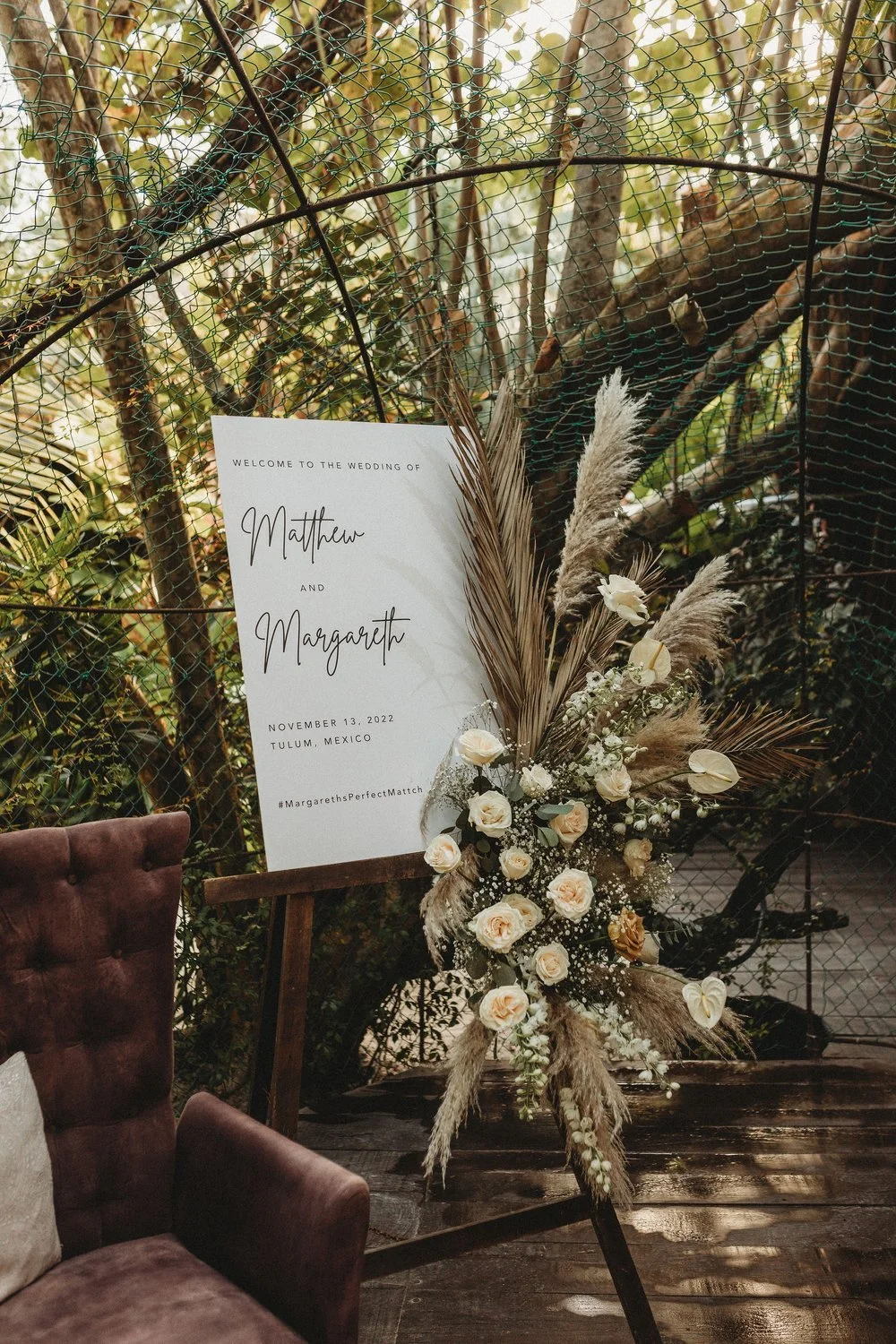 Wedding welcome sign on a stand with floral arrangement of white roses, dried pampas grass, and other flowers, set outdoors among trees with greenery.