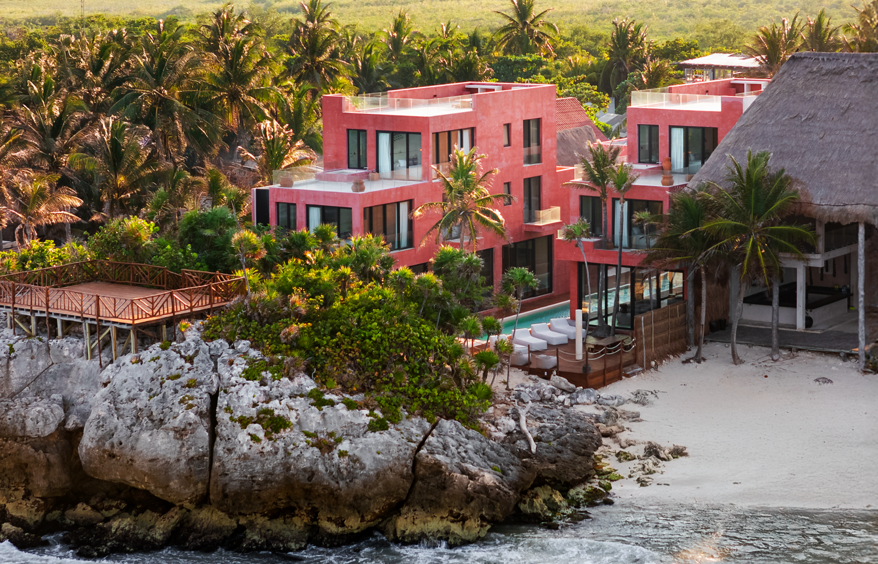 Beachfront resort with pink modern buildings, surrounded by tropical palm trees, rocks, and sandy beach, with a pool and lounge chairs.