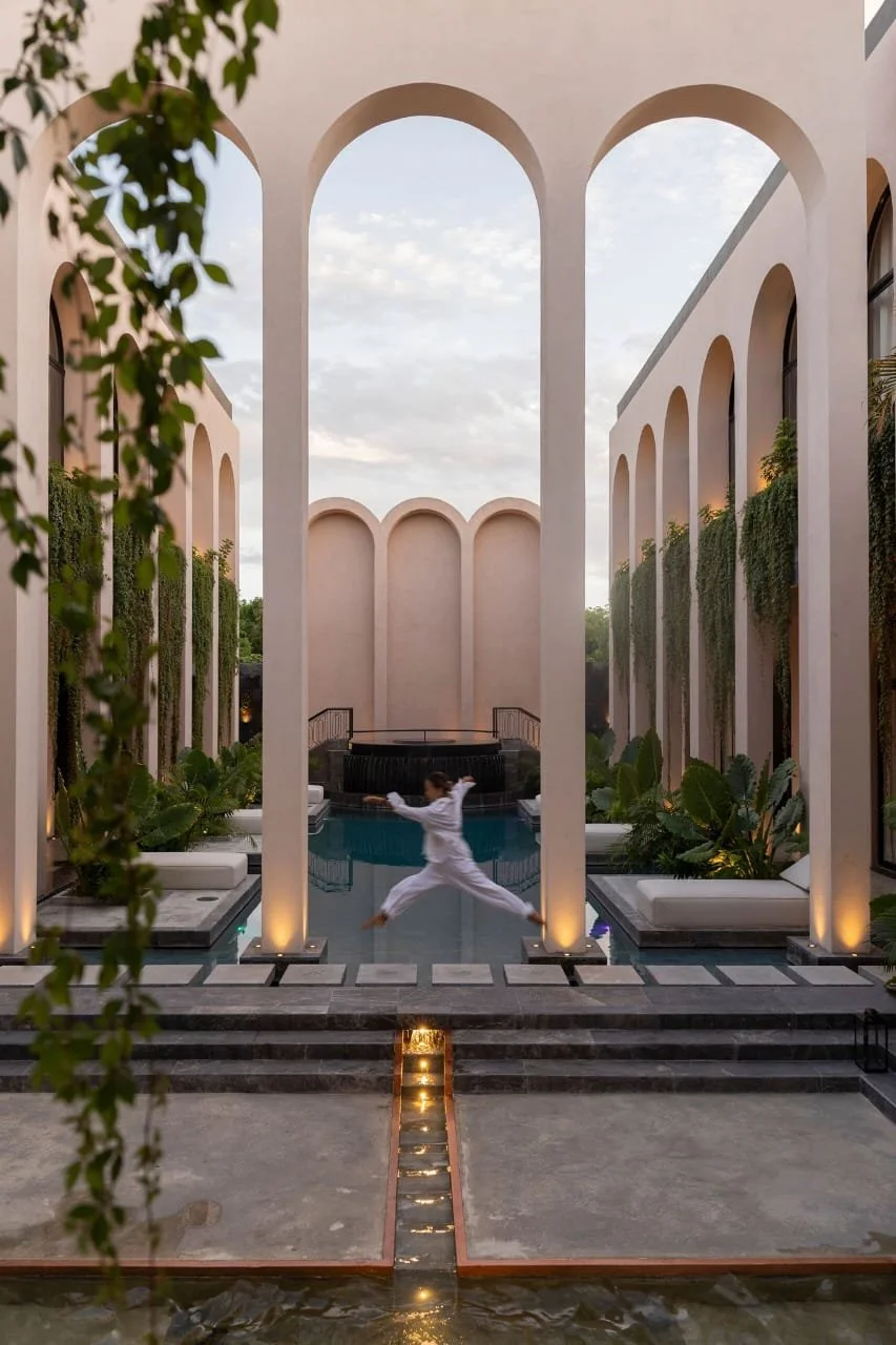 Person in white clothes performing a martial arts move in front of a modern architectural courtyard with tall archways, a pool, and lush greenery.