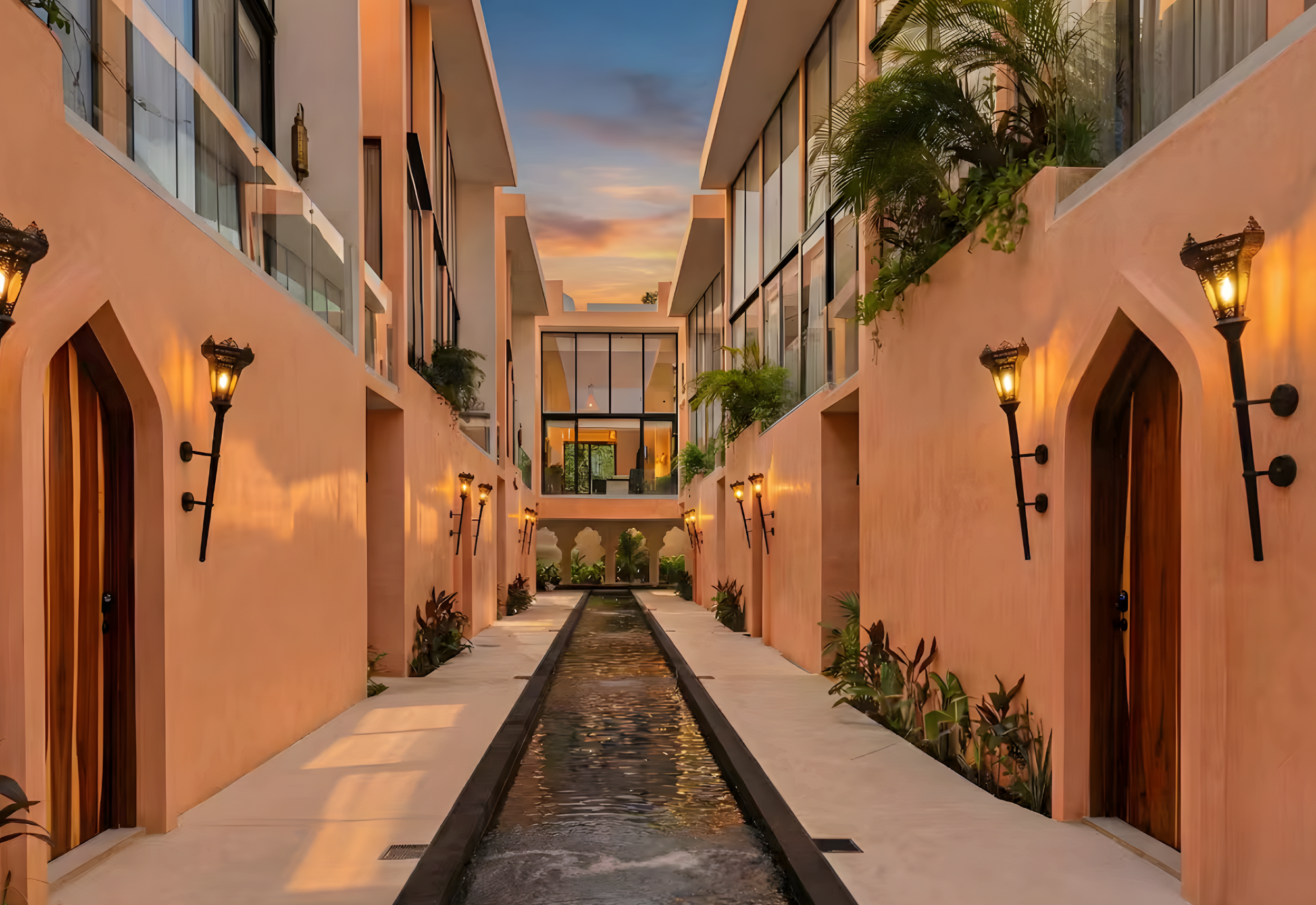 Exterior view of a modern apartment complex courtyard at sunset with a water feature, adjacent walkway, arched doorways, and ambient lighting.