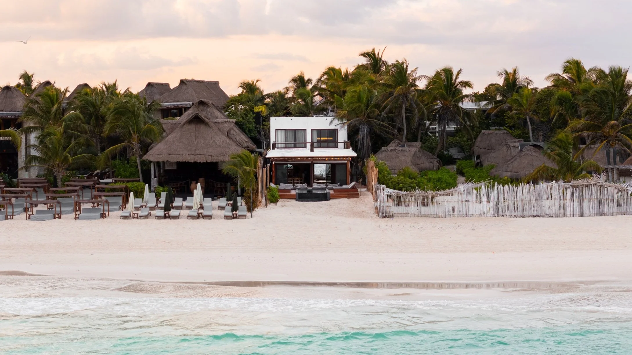Beachfront resort with sandy shore, lounge chairs, and umbrellas, surrounded by palm trees and thatched-roof buildings under a cloudy sky.