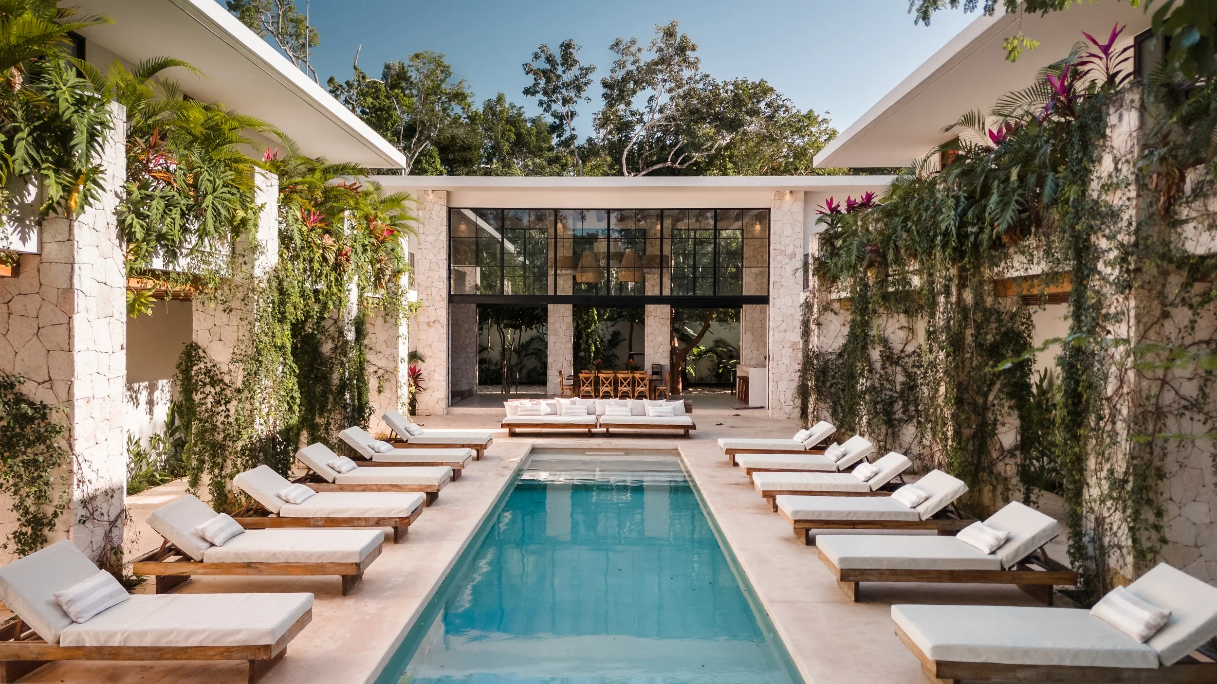 A modern outdoor pool area with white lounge chairs along the sides of a rectangular pool. There are tropical plants climbing stone walls on either side, and a large glass window/door at the end of the pool area showing an indoor space. Trees and a blue sky are visible above.