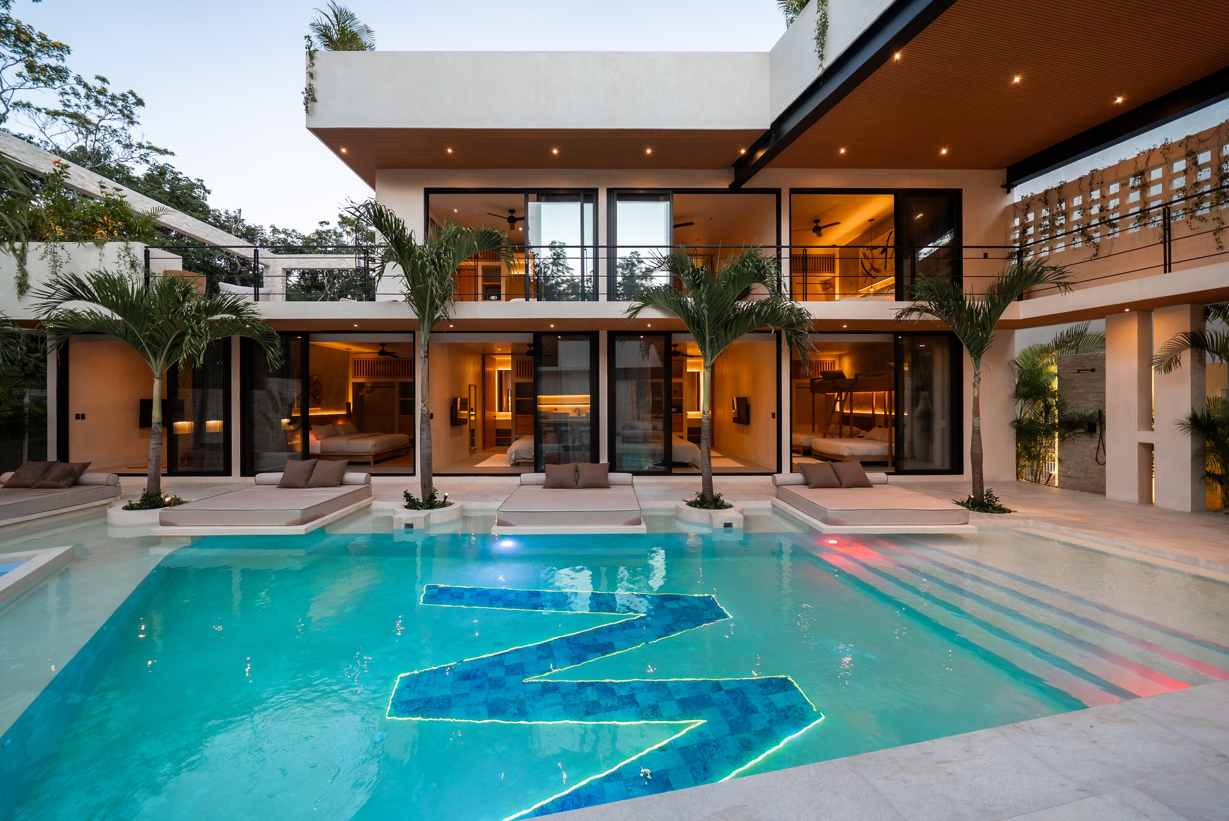 Modern house with large glass doors and an outdoor pool with built-in steps and colorful lighting, surrounded by lounge chairs and palm trees, during dusk.