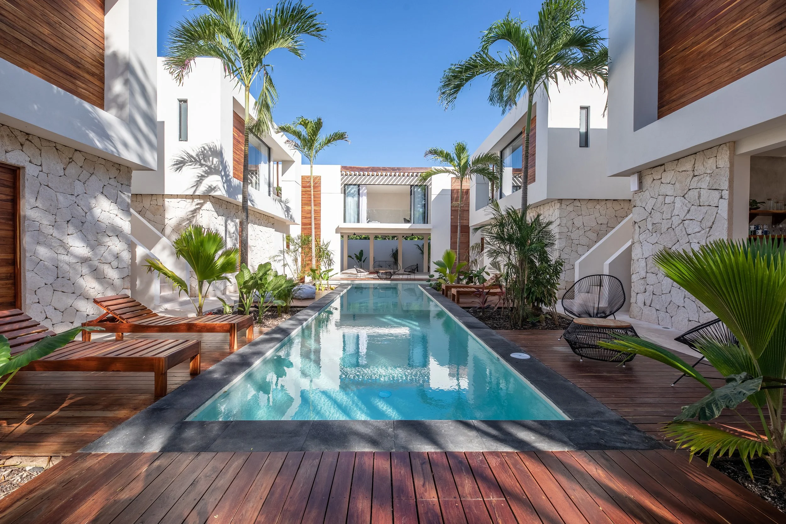 A modern outdoor pool area with a narrow rectangular pool, surrounded by wooden decking and tropical plants. Tall palm trees are positioned around the pool, with white walls and windows of a contemporary building in the background, under a clear blue sky.