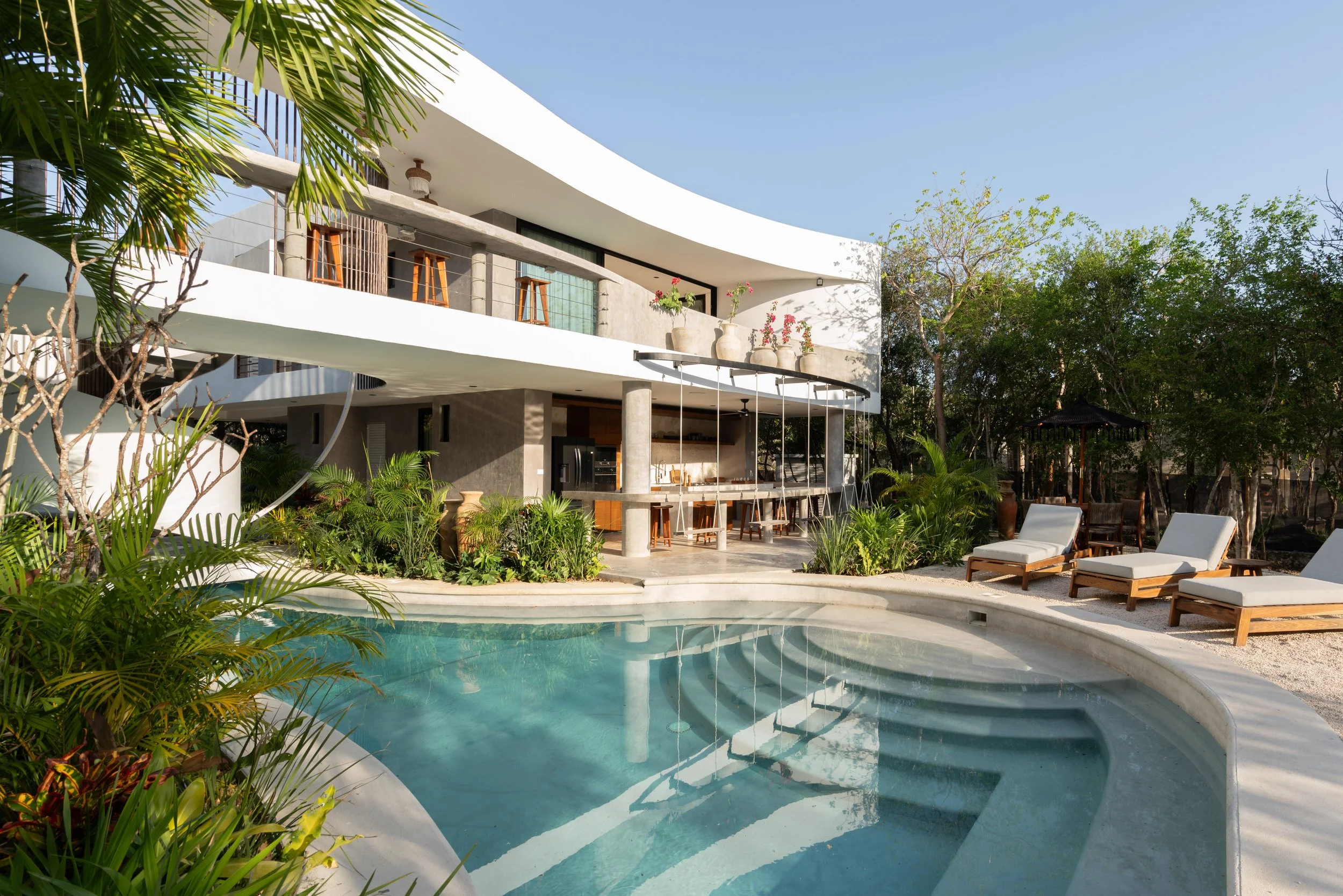 Modern house with outdoor pool, balcony with bar stools, potted plants, and outdoor lounge chairs, surrounded by greenery and trees under a clear blue sky.