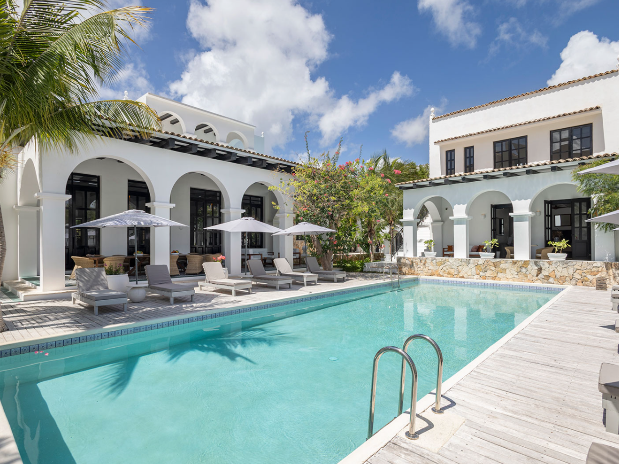 A luxurious outdoor swimming pool surrounded by white Mediterranean-style buildings with arches, black-framed windows, and terracotta roof tiles. There are poolside lounge chairs with white cushions, several white patio umbrellas, and tropical trees and plants, all under a bright blue sky with a few clouds.