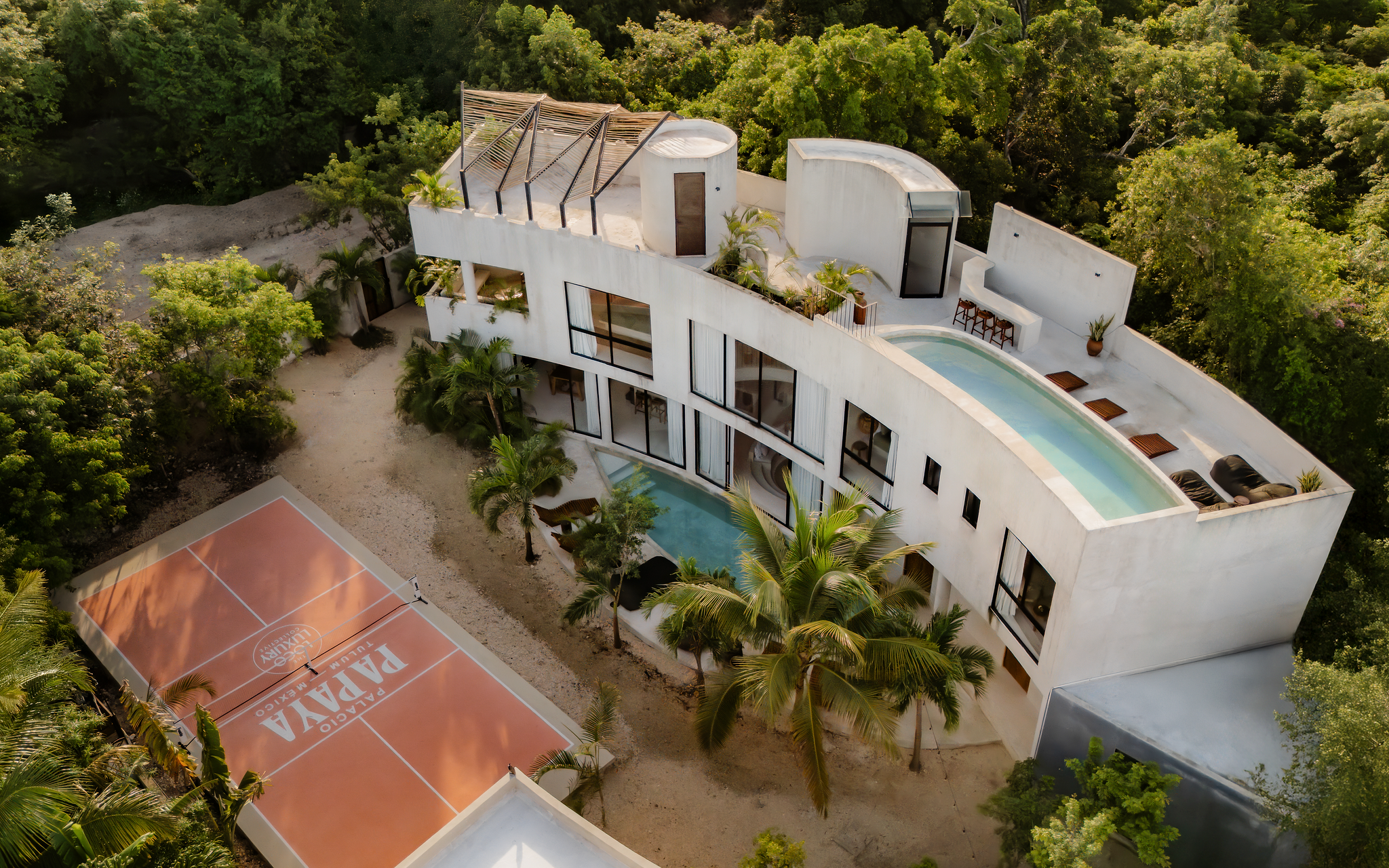 An aerial view of a modern white house surrounded by lush green trees. The house features large windows, a pool, and a rooftop patio with sun loungers. There is also a tennis court nearby.