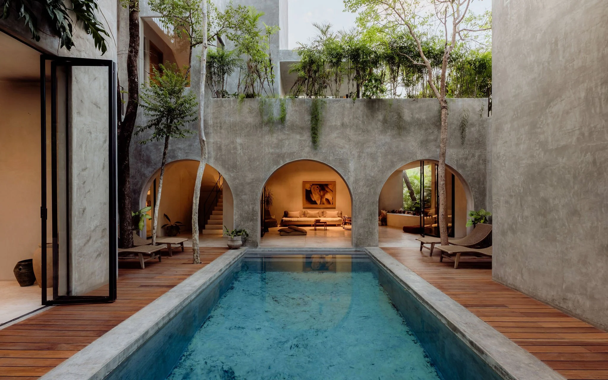 A modern indoor courtyard with a rectangular swimming pool, surrounded by wooden decking, featuring trees and plants in planters, with arched openings and a seating area visible through the arches.