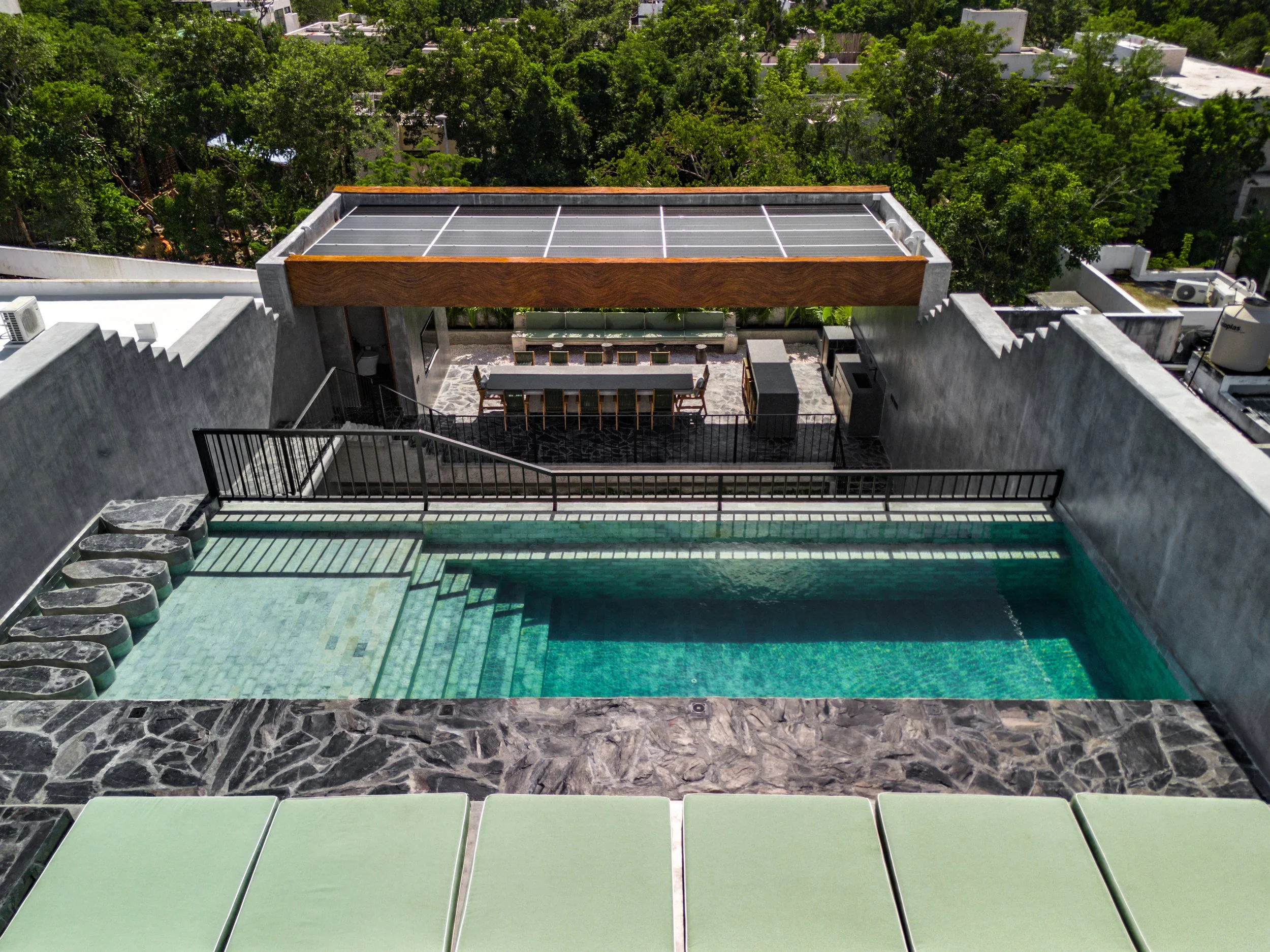 A rooftop pool area with steps leading into the pool, surrounded by stone tiles, and a terrace with a long dining table and chairs. Above the terrace is an architectural wooden beam with a solar panel installation on top. In the background, lush green trees and neighboring rooftops are visible.
