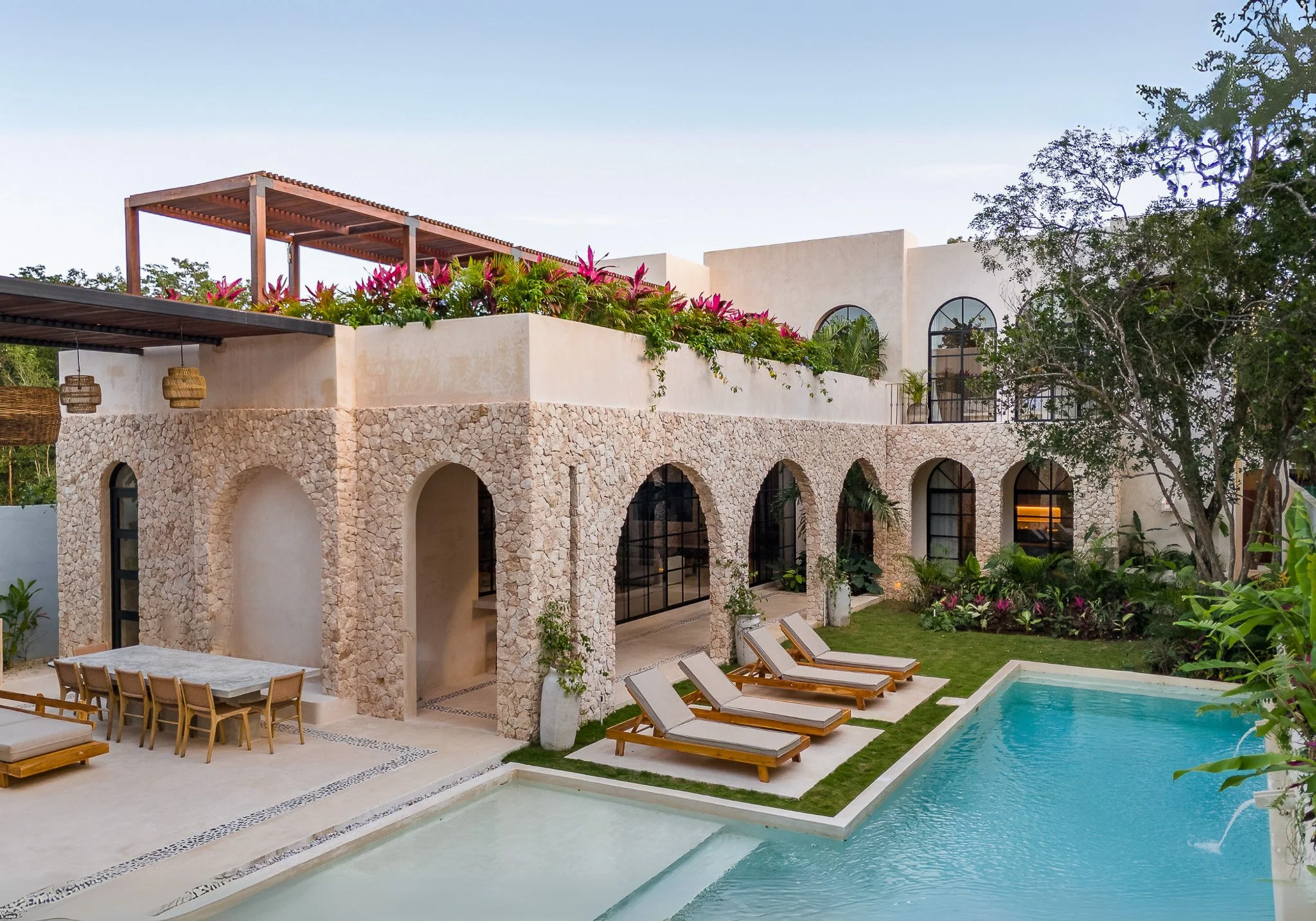 A modern house with stone arches on the ground floor, a white stucco upper level, large arched windows, and a rooftop terrace with pink flowering plants, a wooden pergola, and outdoor furniture beside a swimming pool and lounge chairs.
