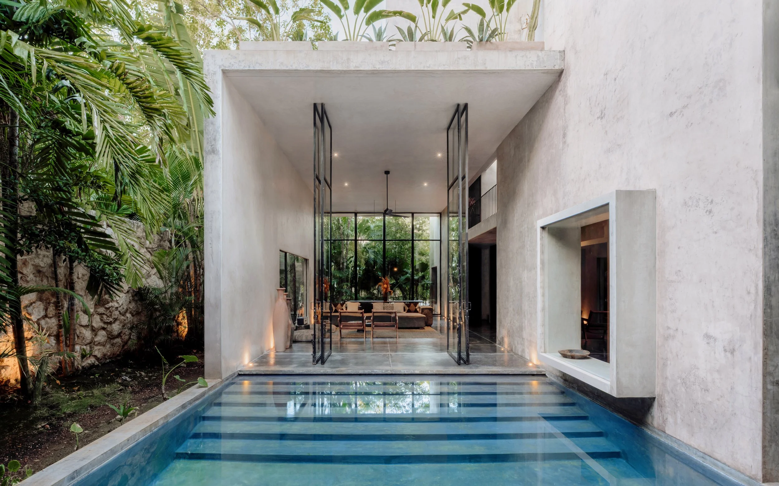Modern house with open living area, large glass doors, and a pool in the foreground, surrounded by greenery.
