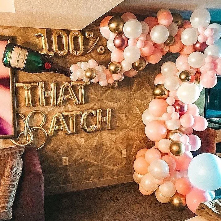 Decorative balloon arrangement in pink, white, and gold, with a large gold '100% that bachelorette' balloon and a bottle-shaped balloon. Gold letter balloons spell out 'That Bitch' on a wood-patterned wall.