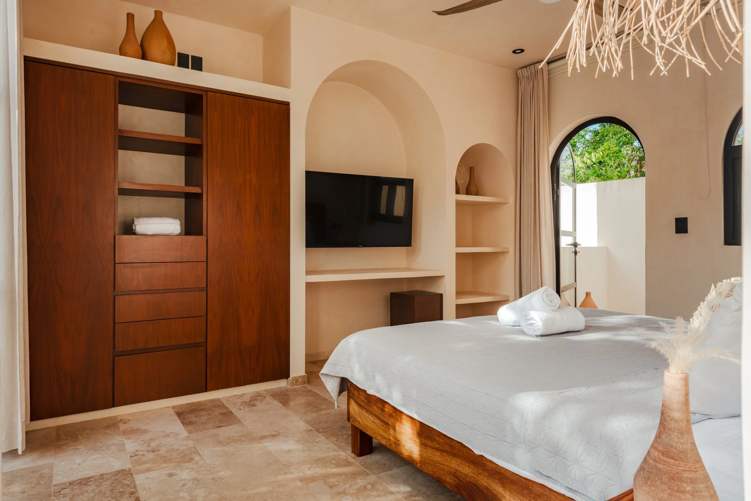A bedroom with a bed, white linens, and rolled towels on top. There is a wooden wardrobe and a wall-mounted flat-screen TV. An arched window with beige curtains shows green foliage outside. The room has a neutral color palette and a modern, minimalist design.