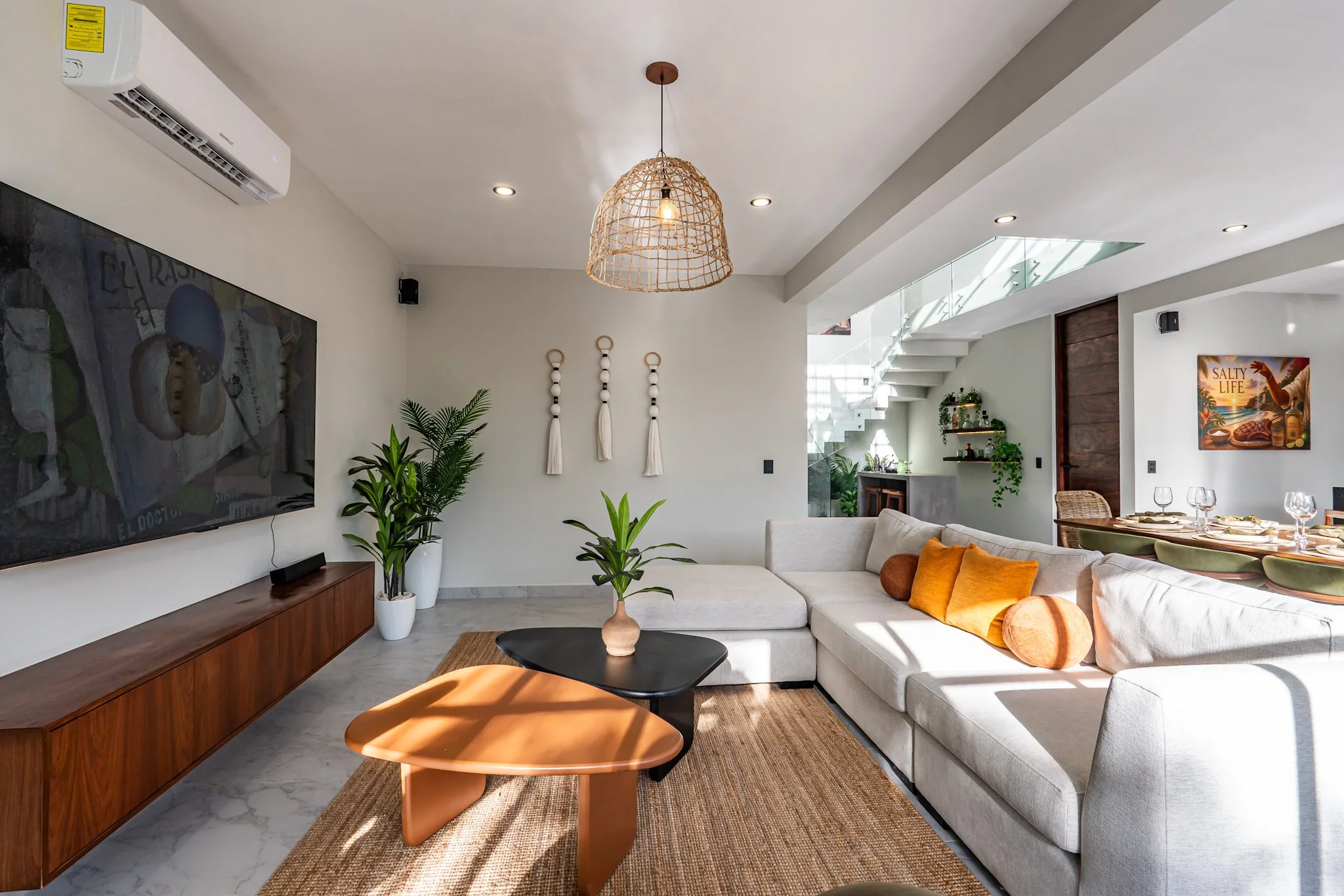 Modern living room with white sofa, orange pillows, black coffee table, wooden entertainment center, wall-mounted TV, indoor plants, and decorative wall hangings. Dining area visible in background with table set with glasses and placemats, stairs leading to upper level, and contemporary lighting fixtures.