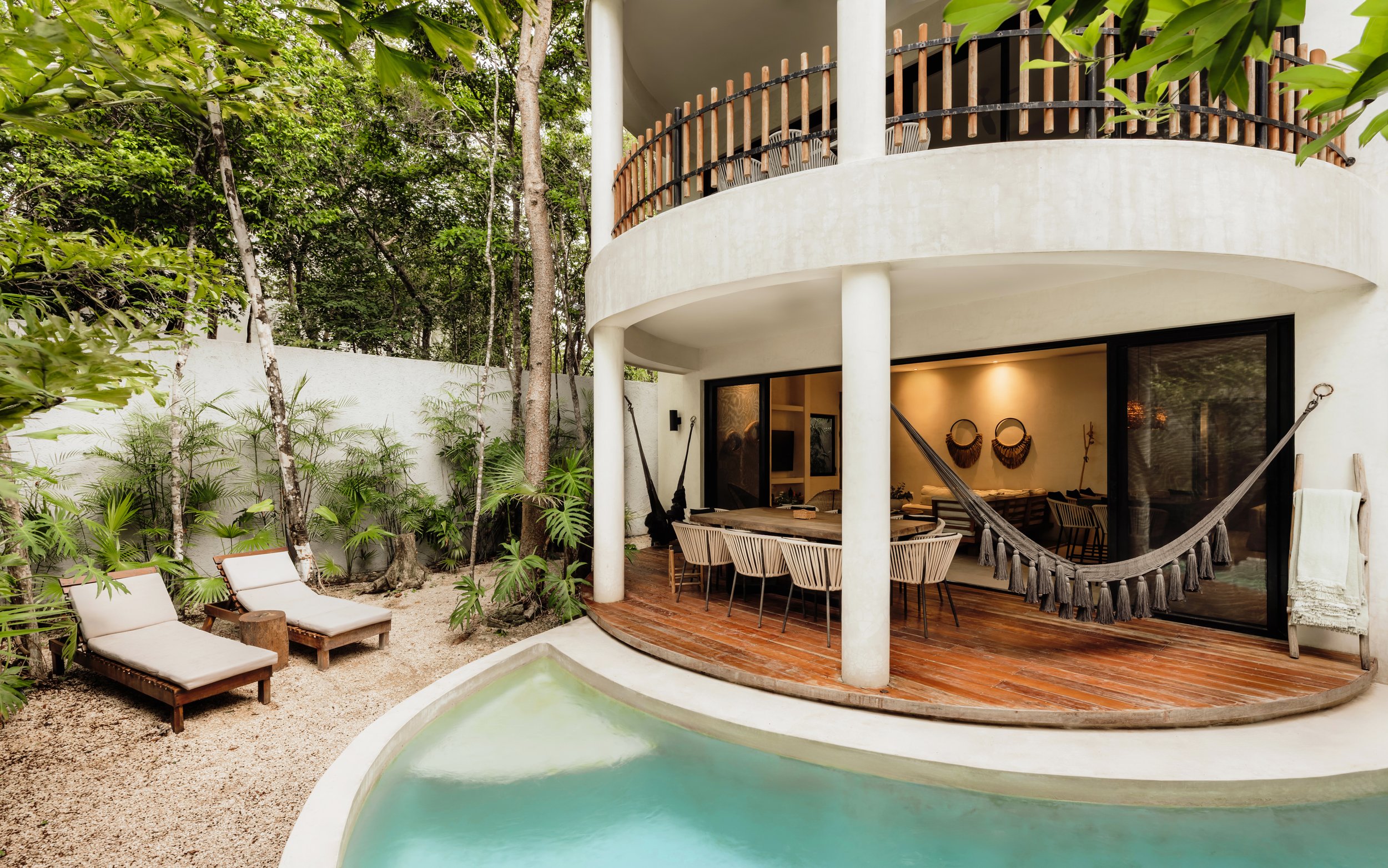 Exterior view of a modern house with a rounded balcony, surrounded by lush green trees and a small outdoor seating area with lounge chairs. The house has large glass doors leading to an indoor living space with tropical decor.