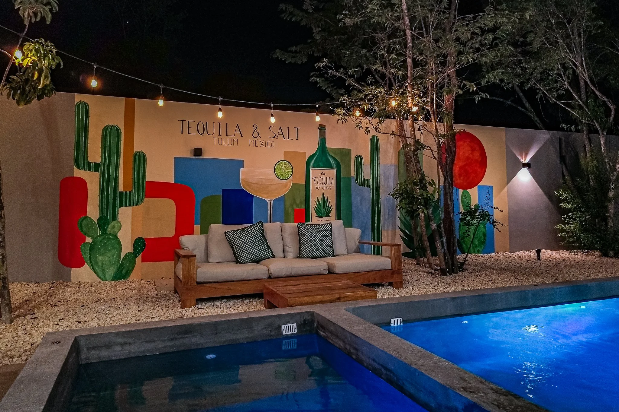 Nighttime outdoor setting featuring a wall with colorful Mexican-themed mural, cactus illustrations, a large bottle graphic, and the text 'Tequila & Salt Tulum Mexico.' There is a wooden sofa with black and white pillows, trees, string lights, and a swimming pool in the foreground.