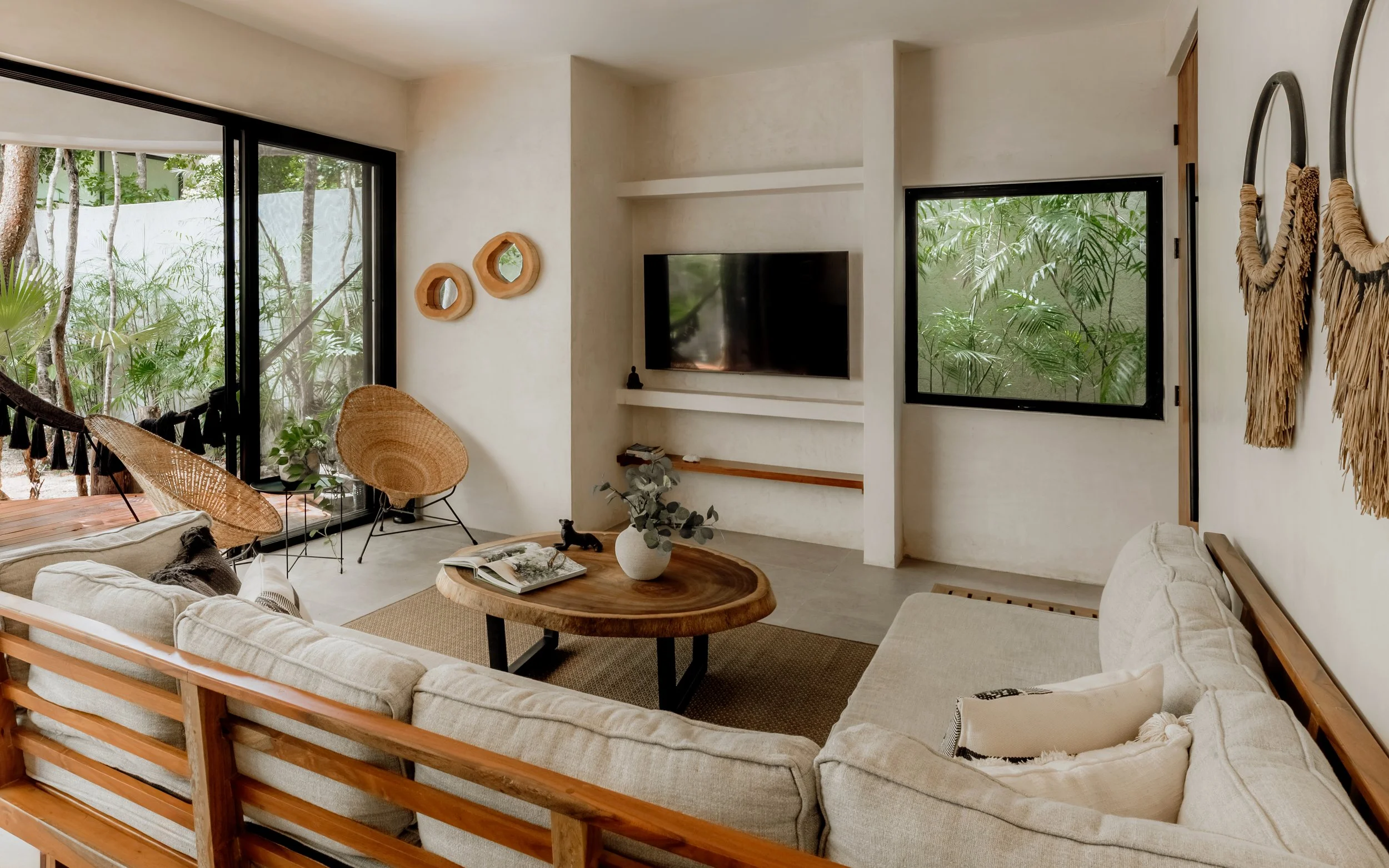 Living room with large sliding glass doors overlooking a lush outdoor garden, beige sectional sofa, and round wooden coffee table with a vase of eucalyptus leaves, wall-mounted flat-screen TV, and decorative wall art.