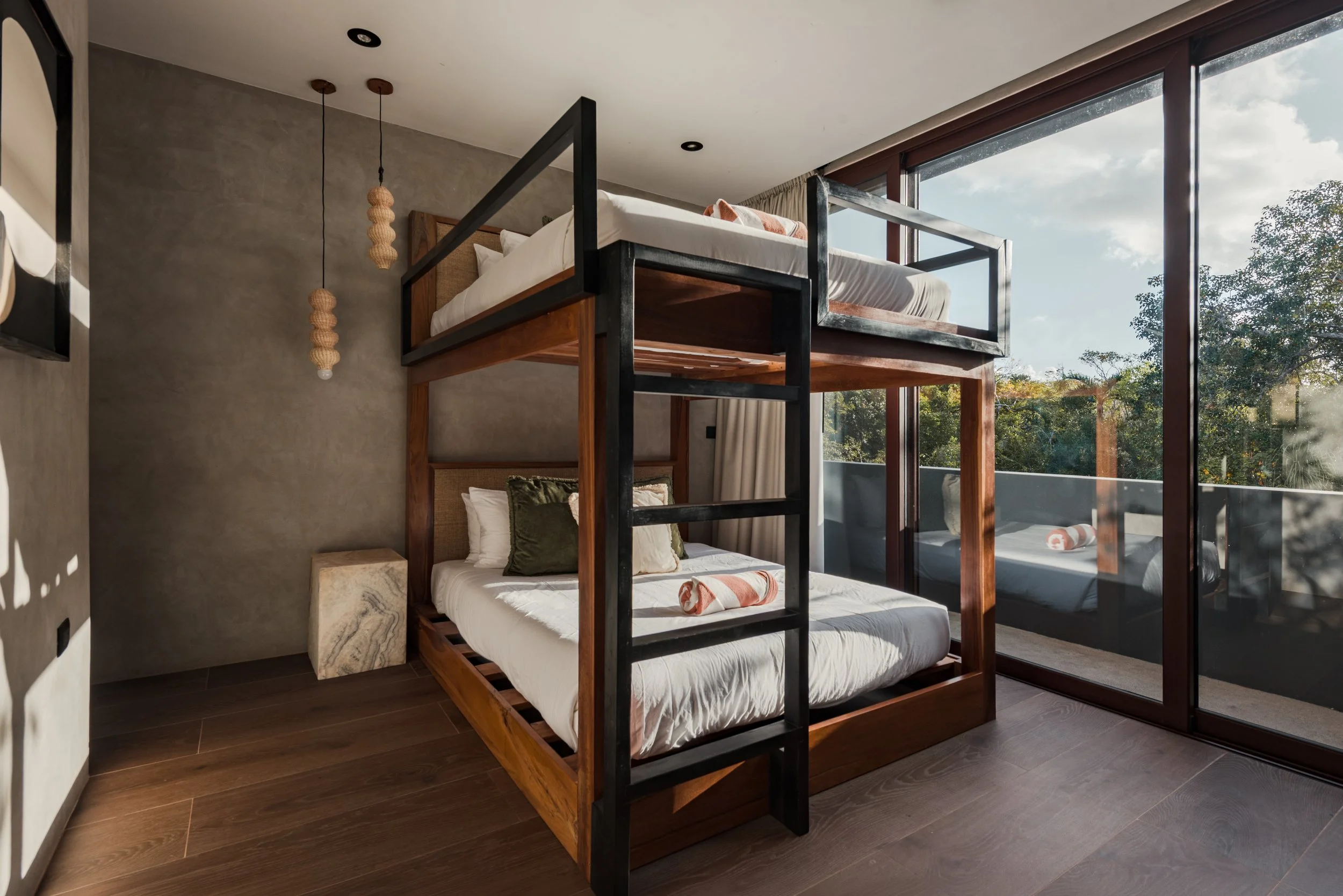 Modern bedroom with a bunk bed near large glass windows showing trees and sky, decorated with neutral tones and wooden accents.