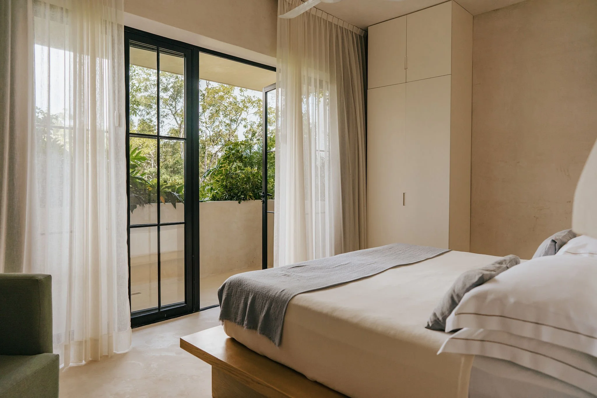 Interior of a bedroom with a bed, sliding glass door, and sheer curtains, overlooking green trees outside.
