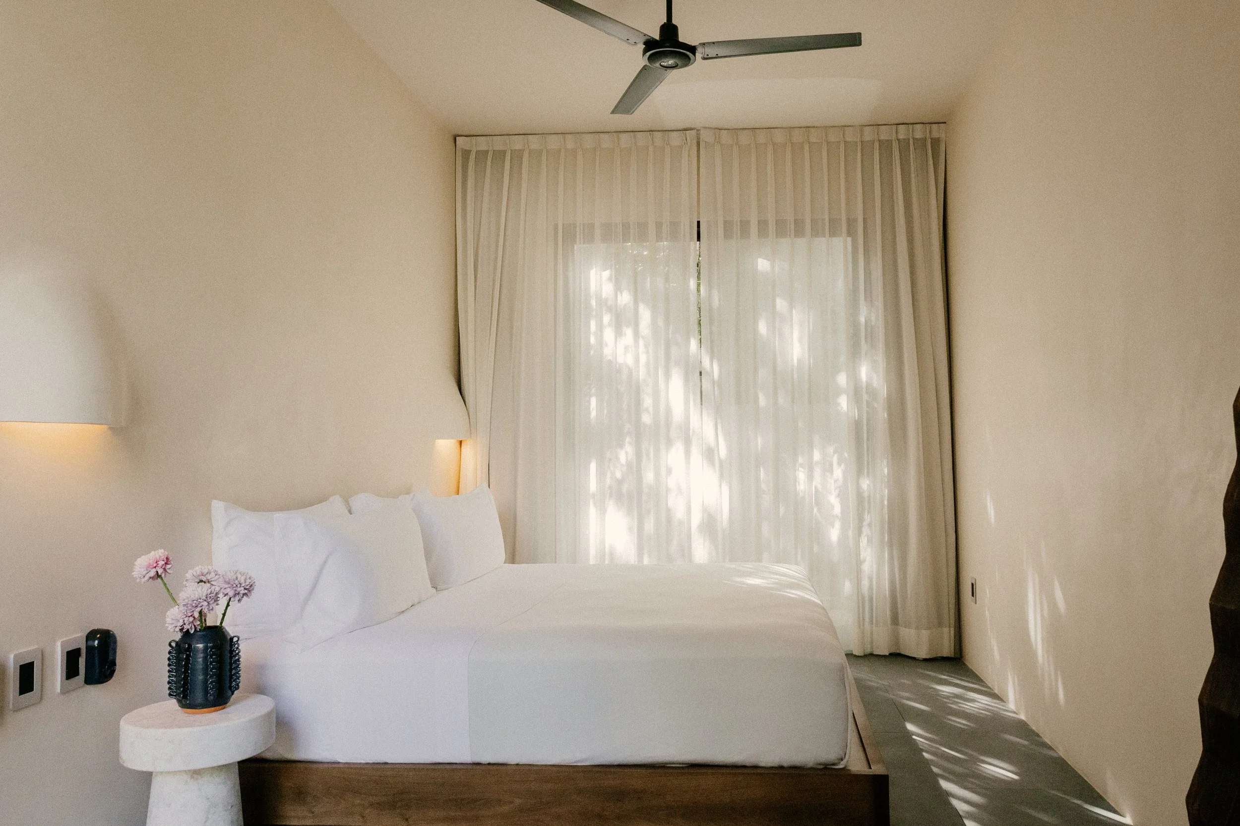 A bedroom with a bed, white bedding and pillows, a small round side table with a flower vase, and a window with sheer curtains. There is a ceiling fan above.