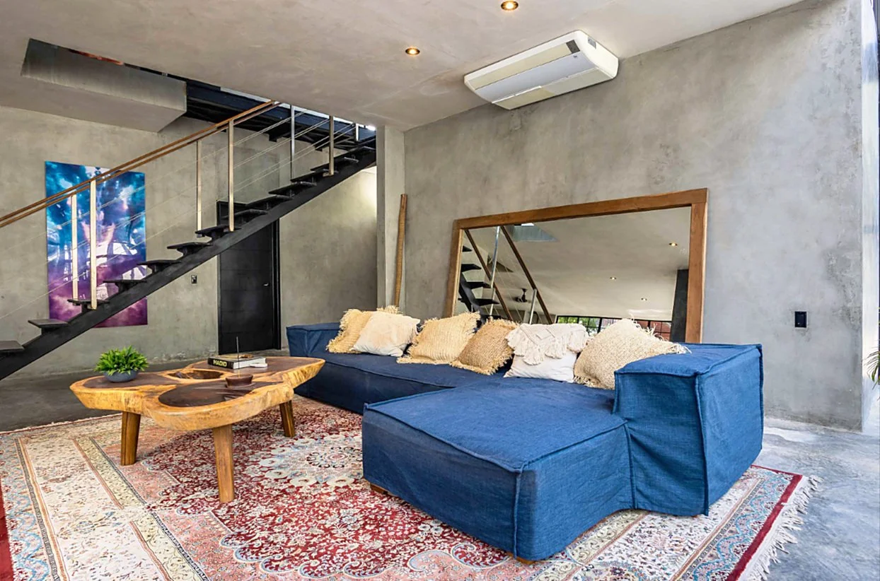 Living room with a large blue sectional sofa, beige pillows, a wooden coffee table with a small green plant, a colorful patterned rug, a large mirror with a wooden frame, a staircase with black steps and a wooden handrail, concrete walls, a ceiling air conditioning unit, and recessed ceiling lights.