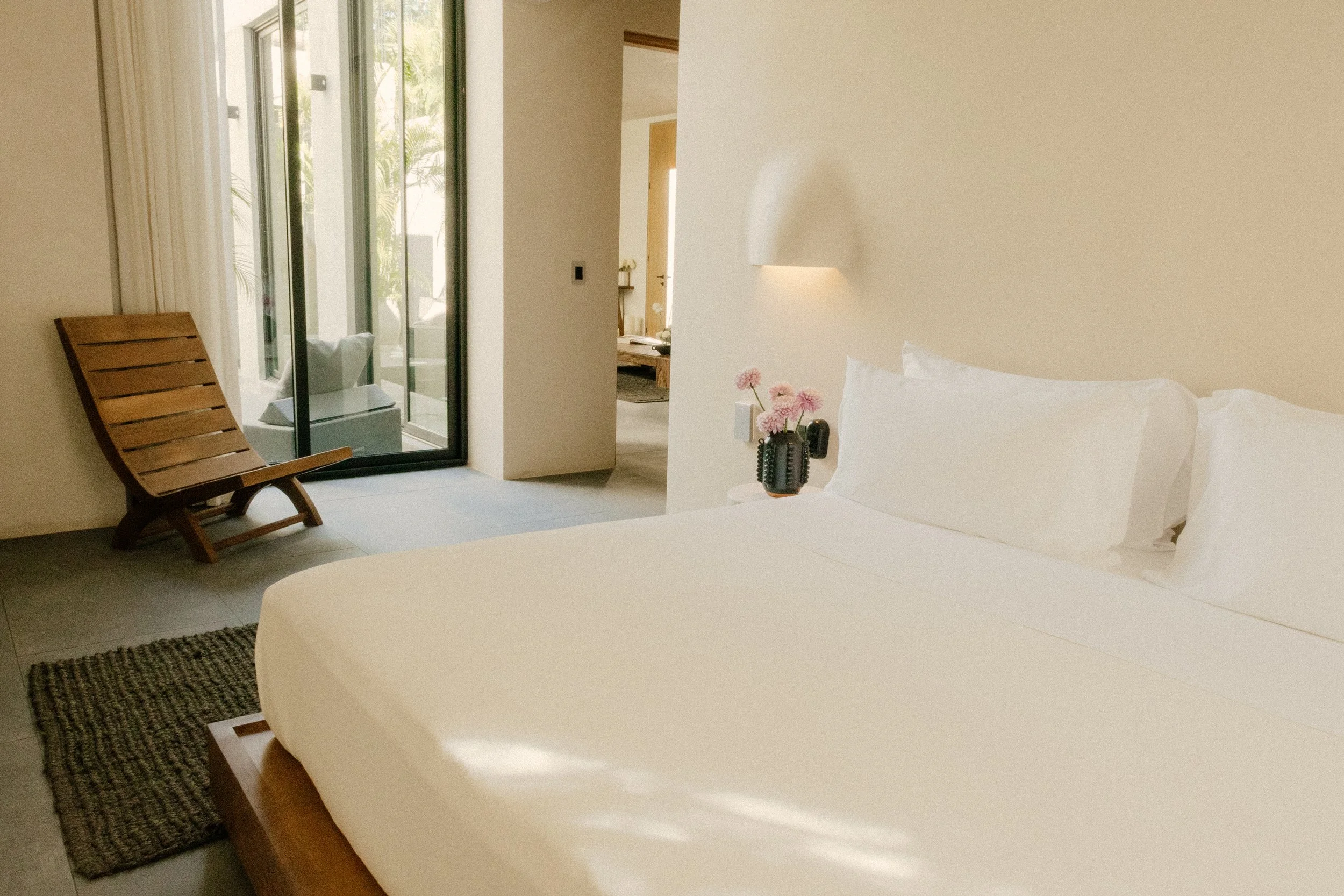 A bedroom with a large bed covered with white linens, a bedside vase with pink flowers, a bedside wall lamp, a black wall switch, a wooden lounge chair, and a sliding glass door leading to a balcony with outdoor seating.