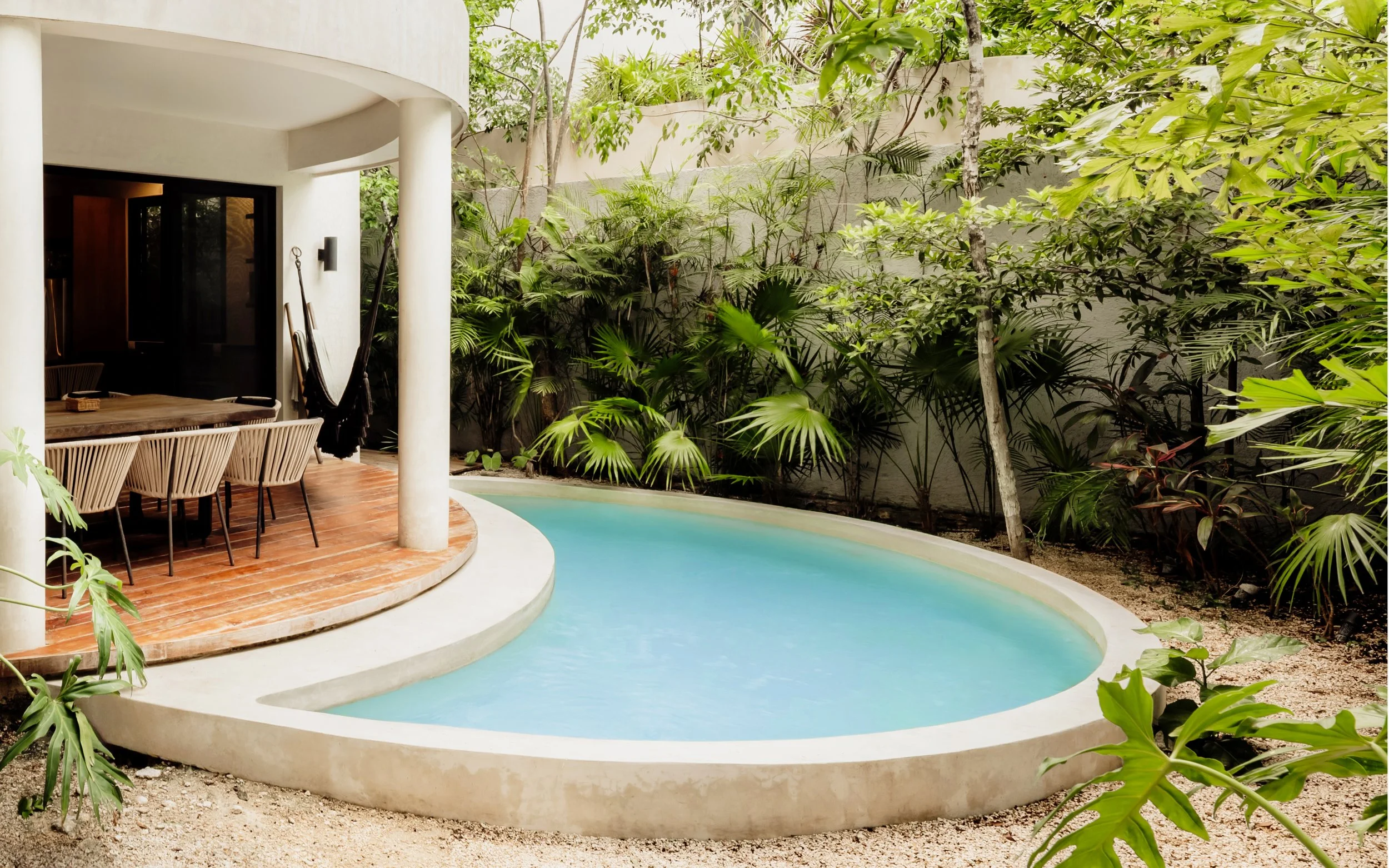 A backyard with a curved swimming pool, a wooden deck, and a white exterior wall. There are lush green plants and trees along the perimeter, creating a lush natural environment.