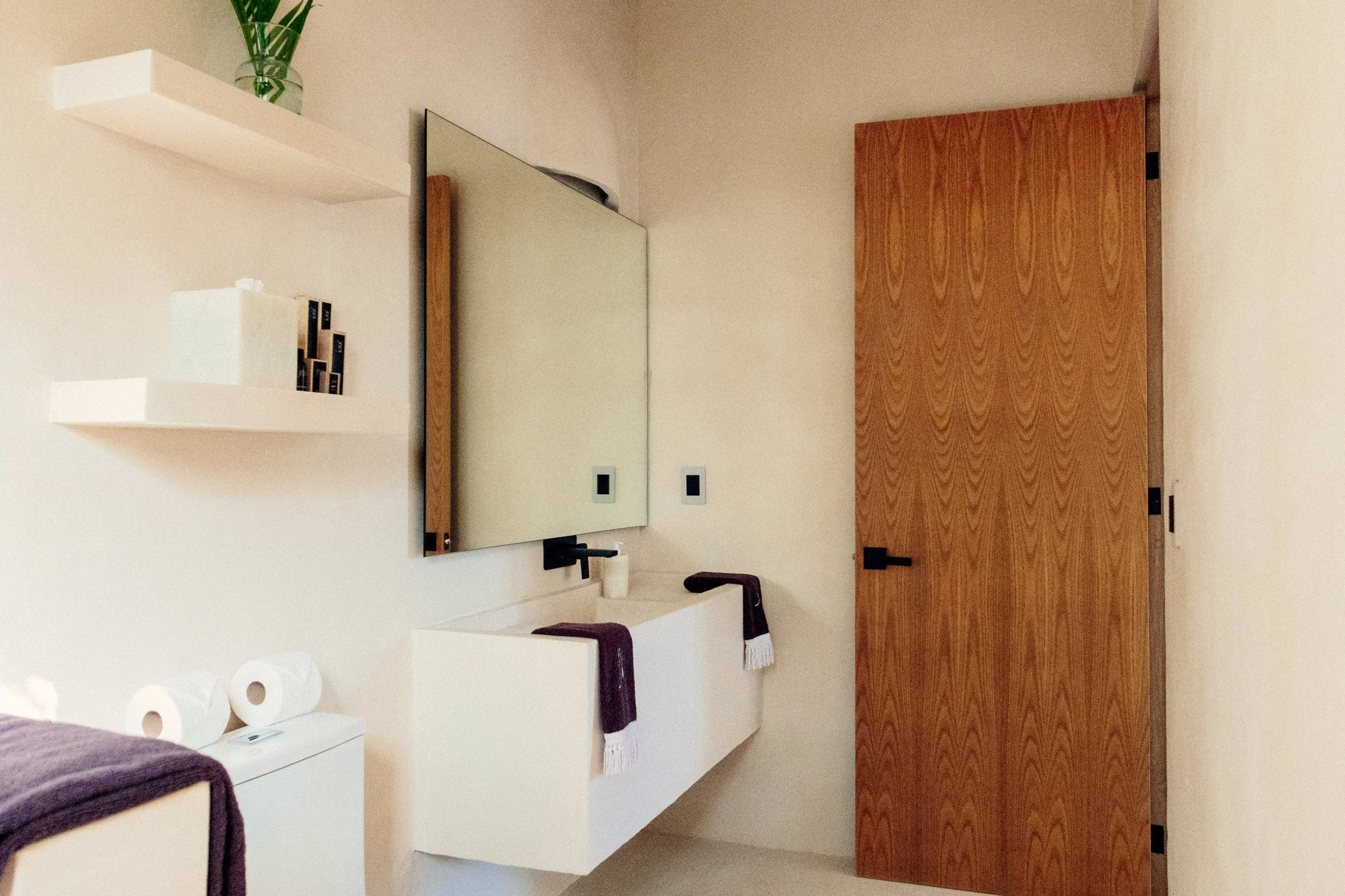 A bedroom with a light-colored wall, two floating white shelves with decorative items, a large rectangular mirror with a wooden frame, a dark-colored door, and a white countertop with a purple towel and paper towels.