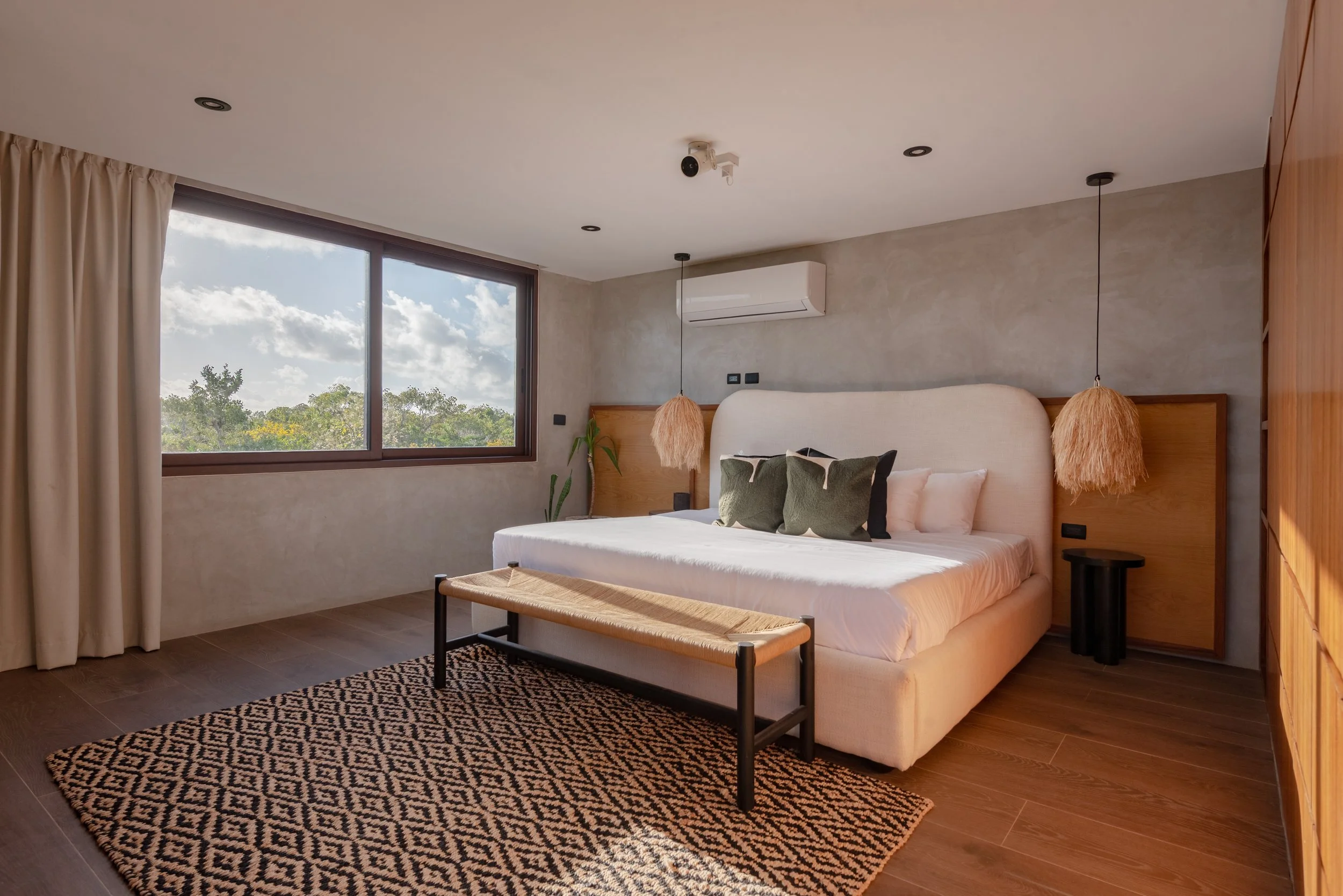 Modern bedroom with large window showing green trees and blue sky, beige curtains, white bed with green throw pillows, black bench, patterned rug, pendant lights, wall-mounted air conditioner, and wooden furniture.