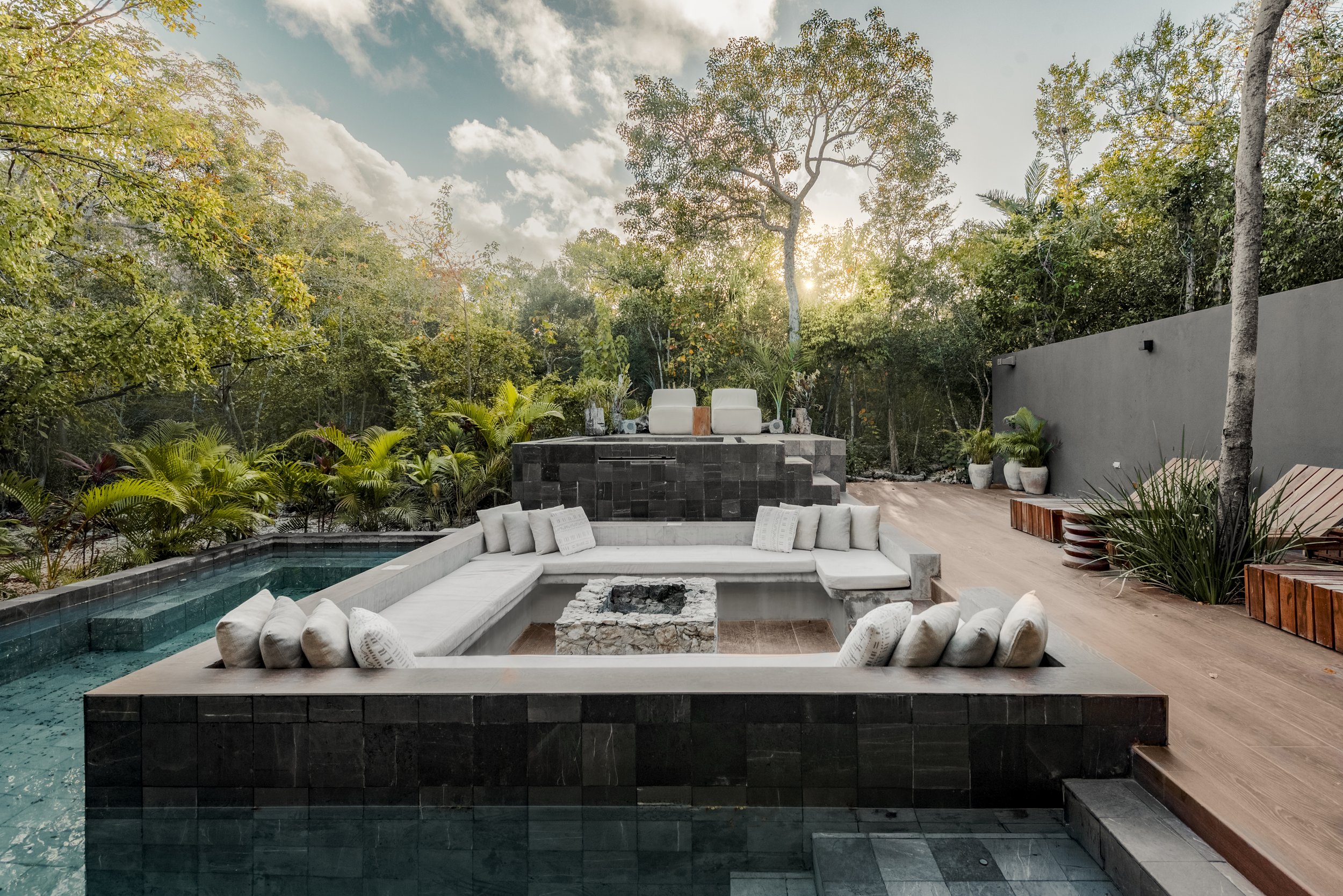 Luxury outdoor patio with a built-in fire pit, adjacent to a swimming pool, surrounded by lush greenery and tall trees, under a partly cloudy sky with the sun setting.