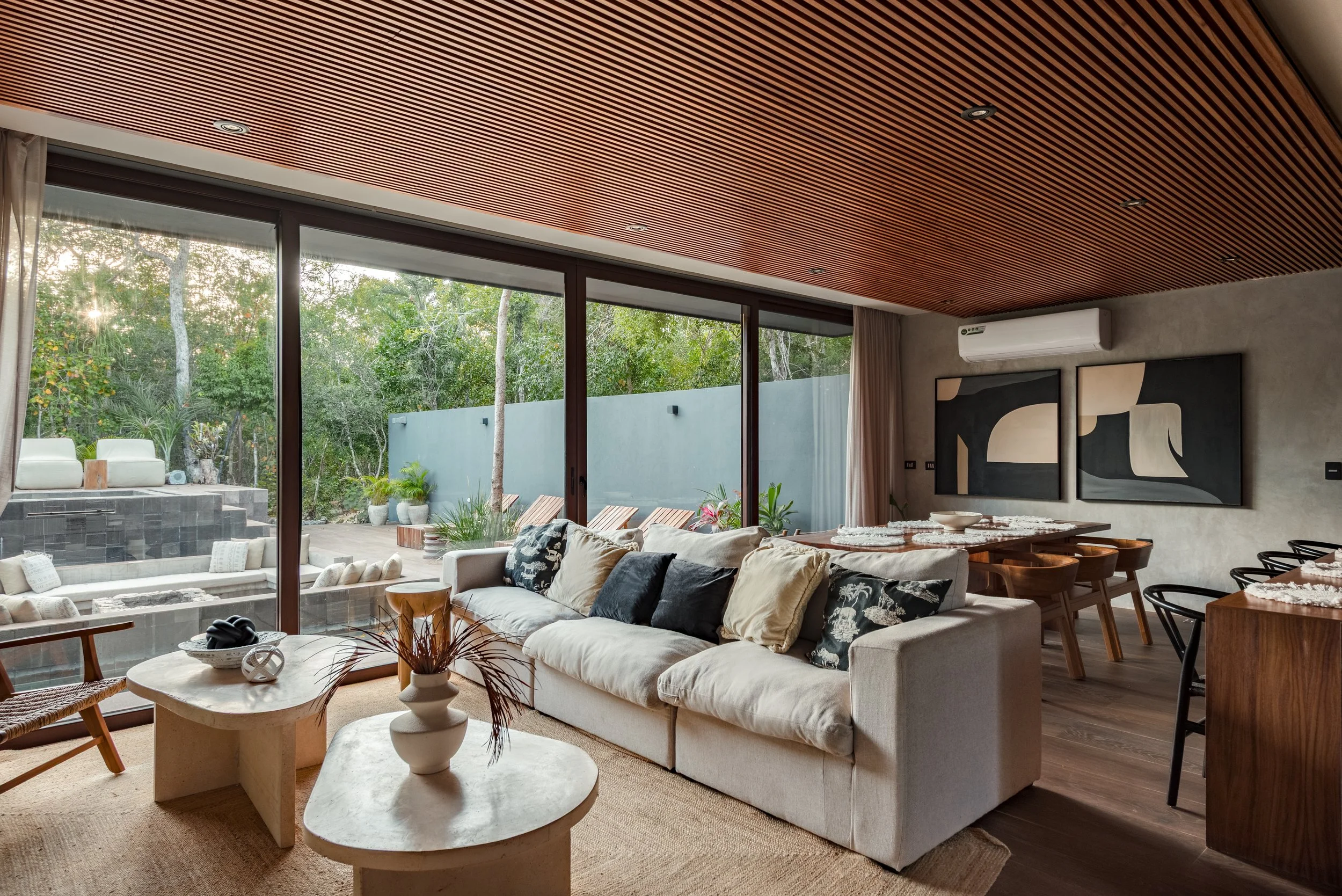 Modern living room with large glass sliding doors showing a lush outdoor garden. Inside, there is a white sofa with decorative pillows, a marble coffee table with a small decorative vase, and a dining area with wooden chairs and a table. The ceiling is wooden with recessed lighting, and there are abstract art pieces on the wall.
