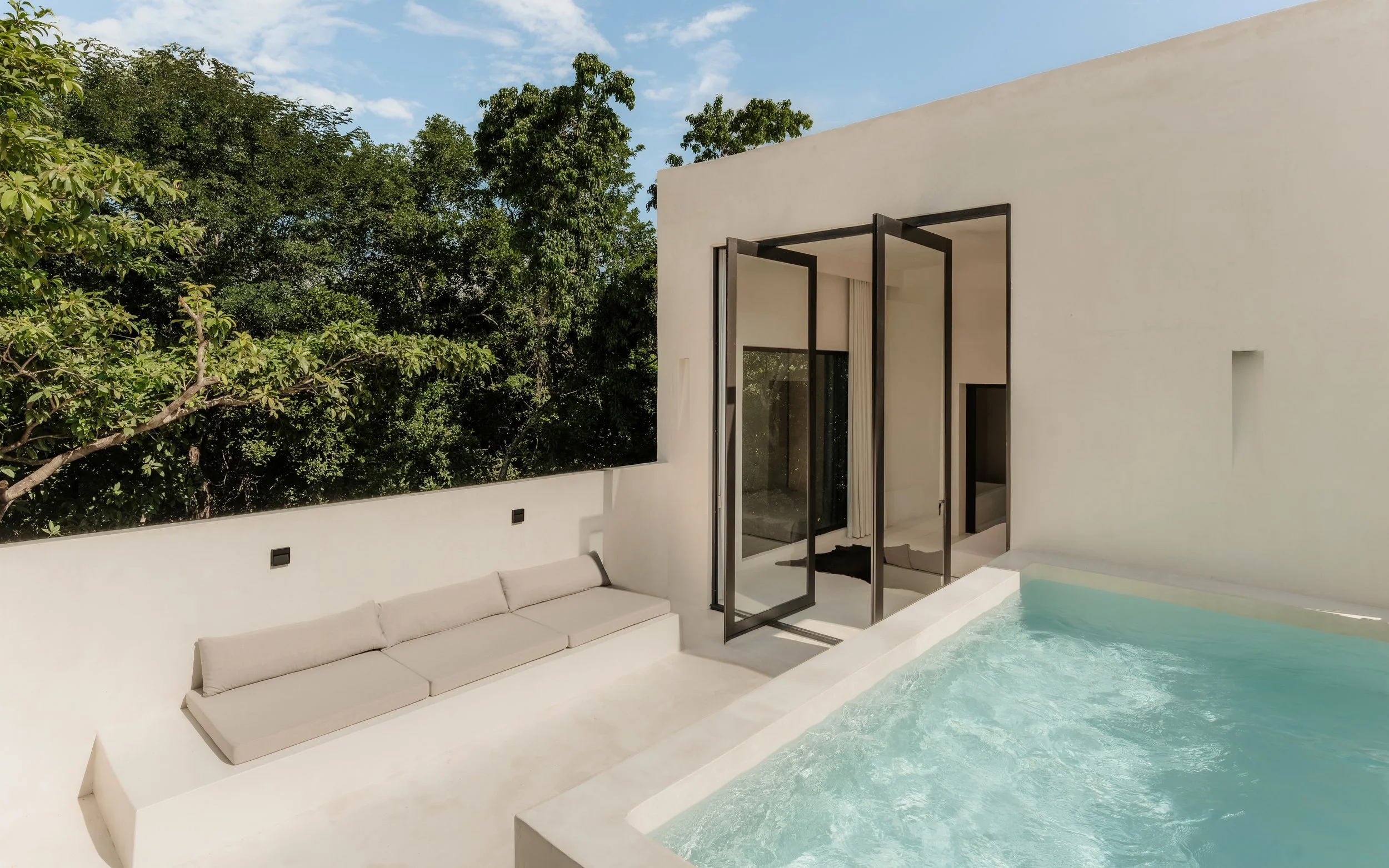 Modern white house with a flat roof, open glass doors, outdoor seating, and a pool, surrounded by green trees and a blue sky.
