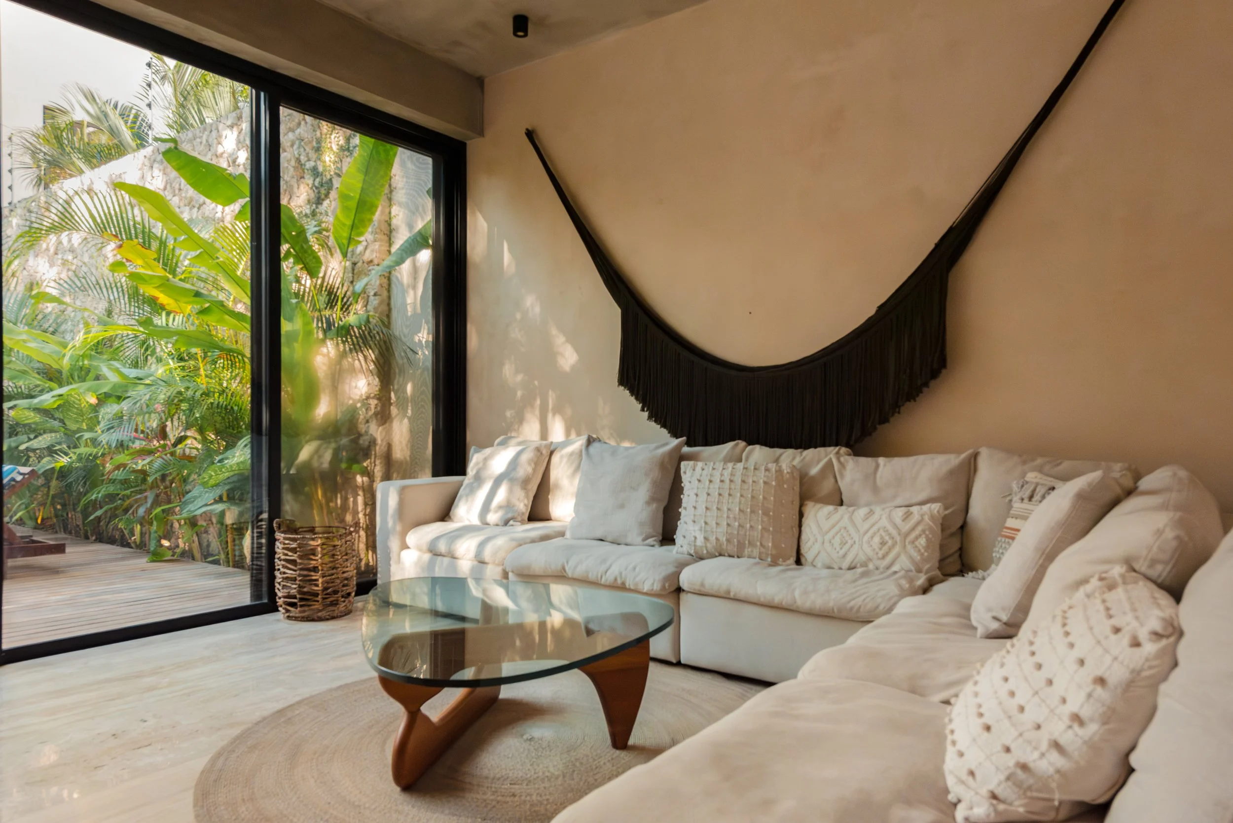 A modern living room with a large beige sofa decorated with white and patterned throw pillows, a glass coffee table with wooden legs, and a view of a lush outdoor garden through a large sliding glass door. Black wall decor is visible above the sofa.