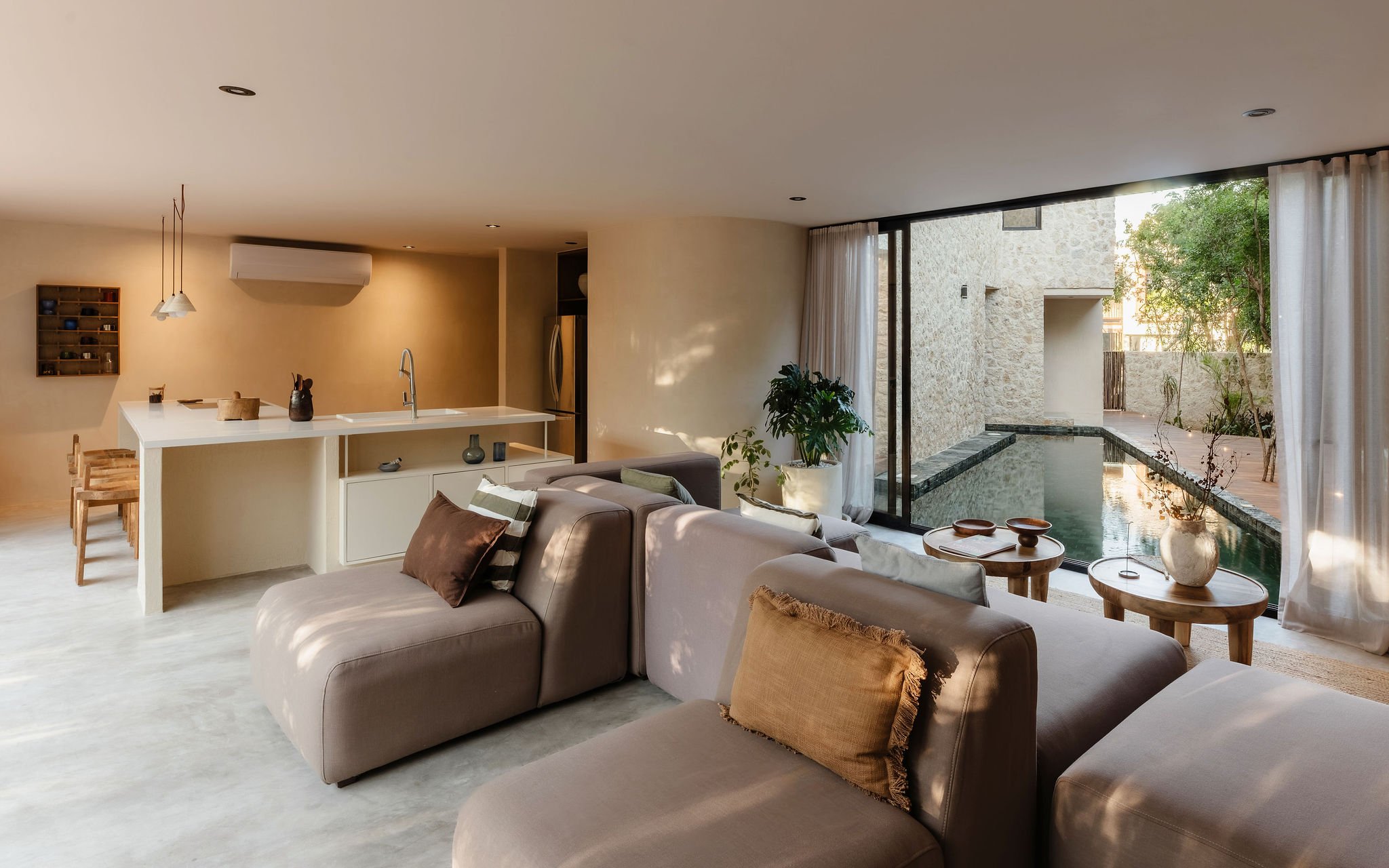 Modern living room featuring neutral-toned sofas with throws, a white island with decorative items, large floor-to-ceiling windows with sheer curtains, and a view of a small outdoor pool area with plants.