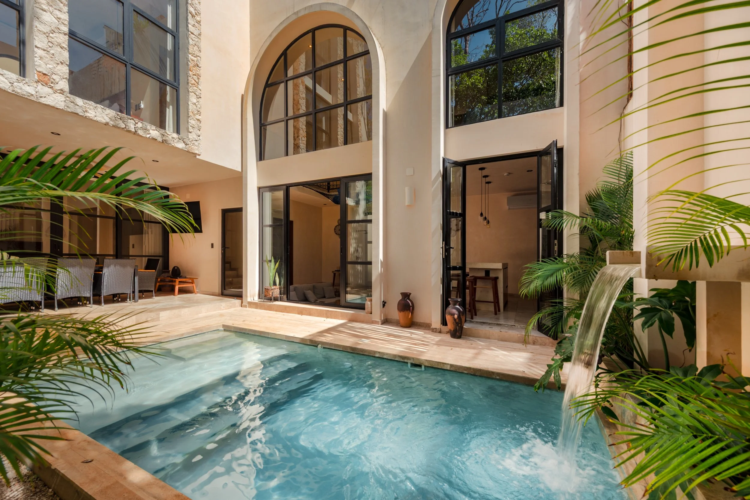 Modern house with large glass windows and doors, a swimming pool, outdoor seating, and lush green plants.