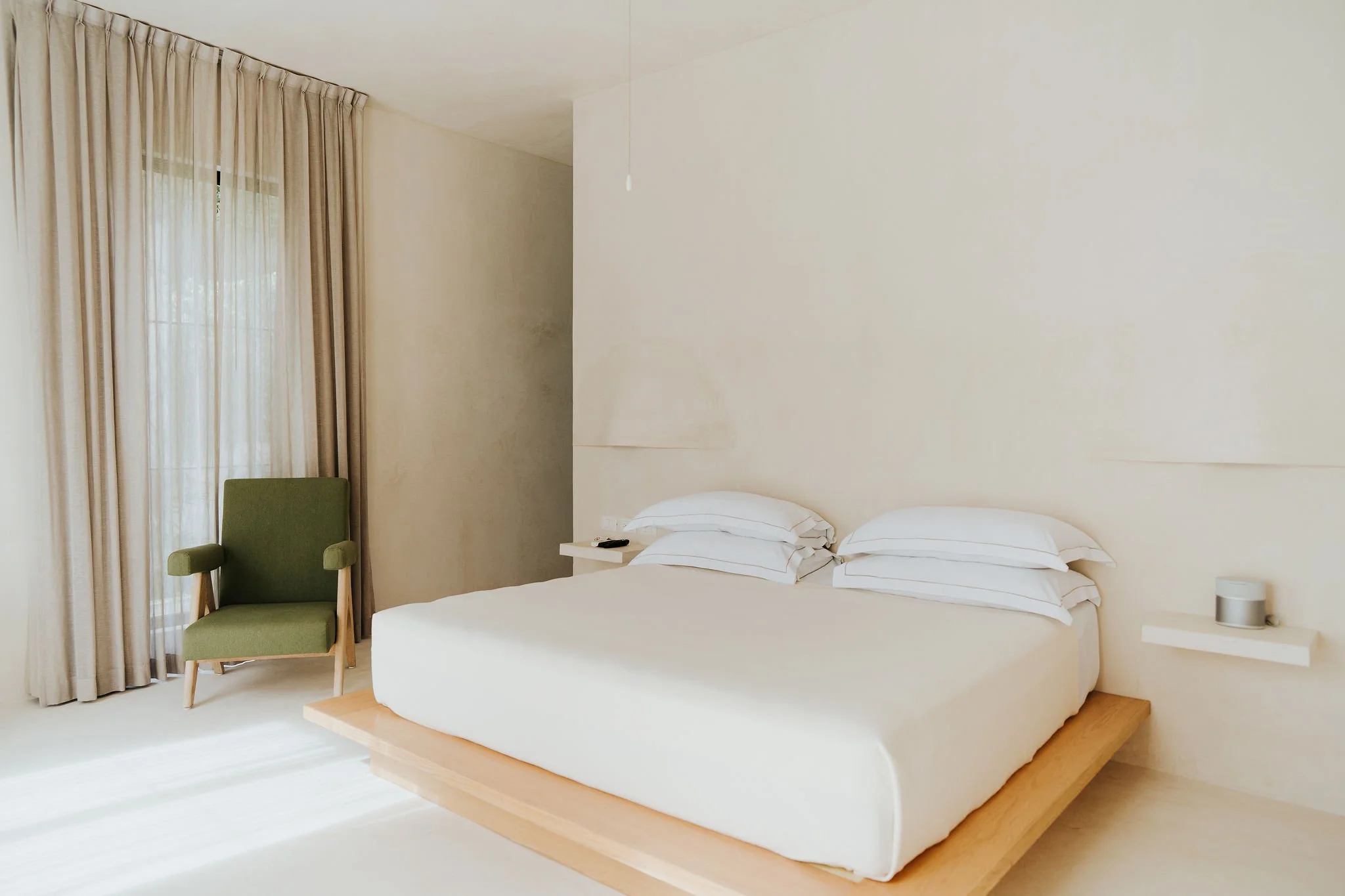 Minimalist bedroom with a low wooden bed frame, white bedding, a bedside table with a remote control, a green armchair, cream curtains, and a window with natural light.