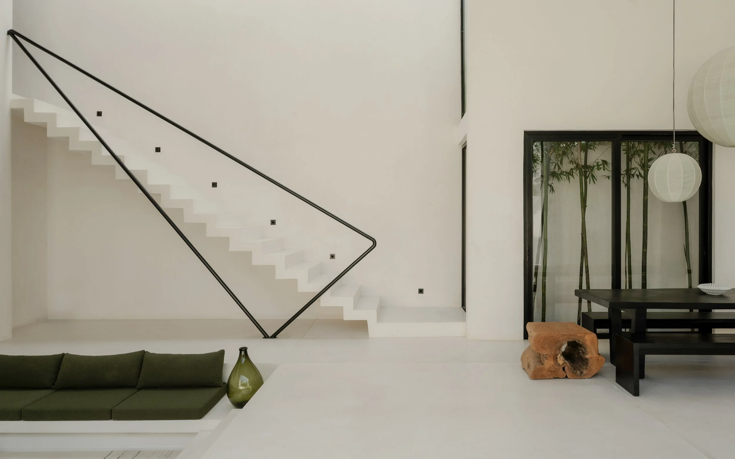 Modern interior with white walls, a staircase with black handrails, a dark wood dining table with a unique wood sculpture, sliding glass doors with bamboo outside, pendant lights, and a green sofa with a green vase.