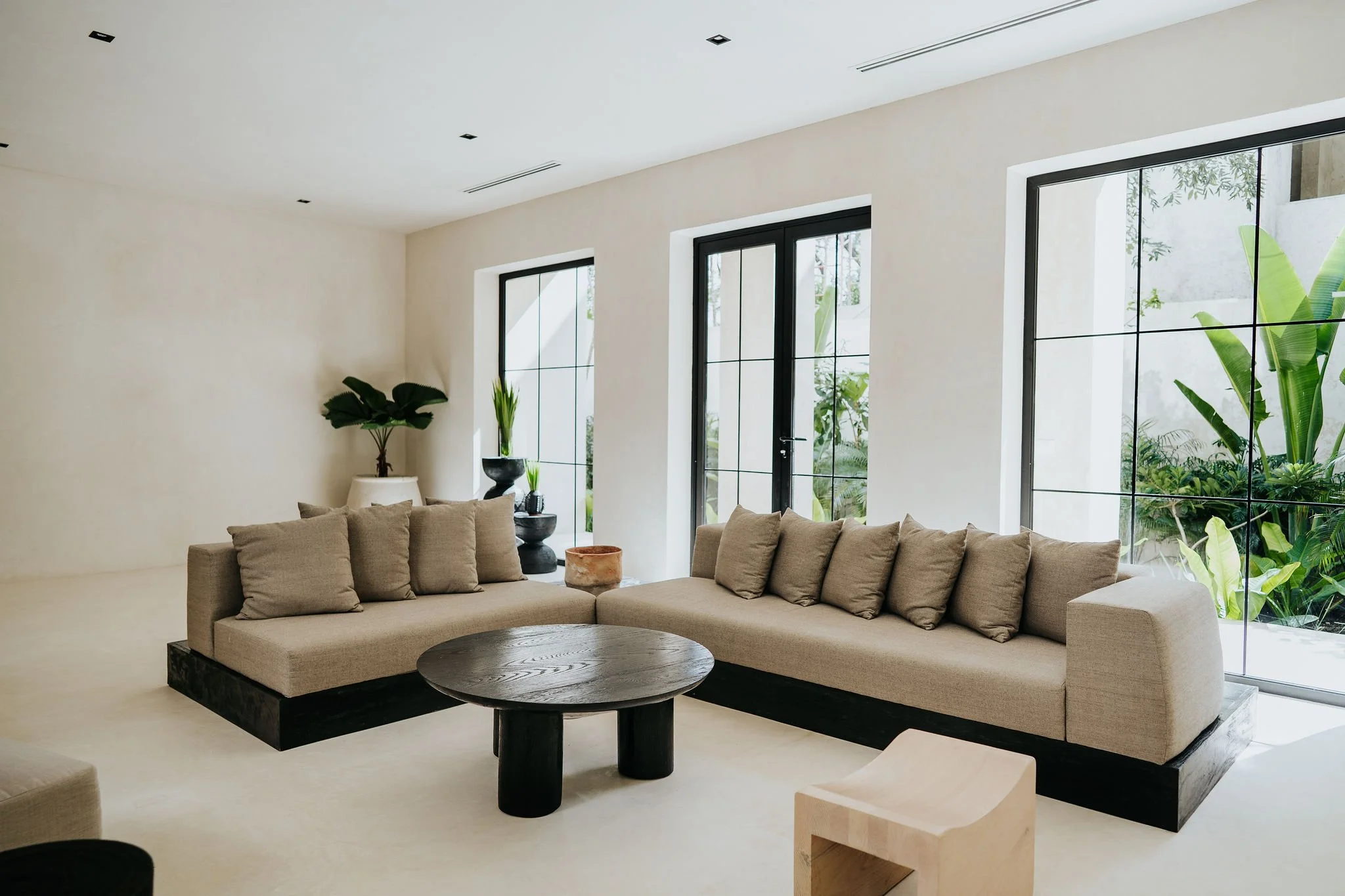 Modern living room with beige sectional sofa, large black-framed windows, and green plants.