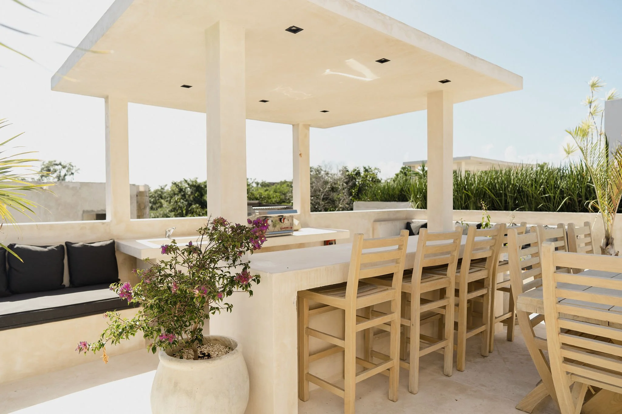 Outdoor patio area with a white ceiling and beige columns, wooden chairs around a large table, a potted plant with pink flowers, and surrounding greenery under a clear sky.